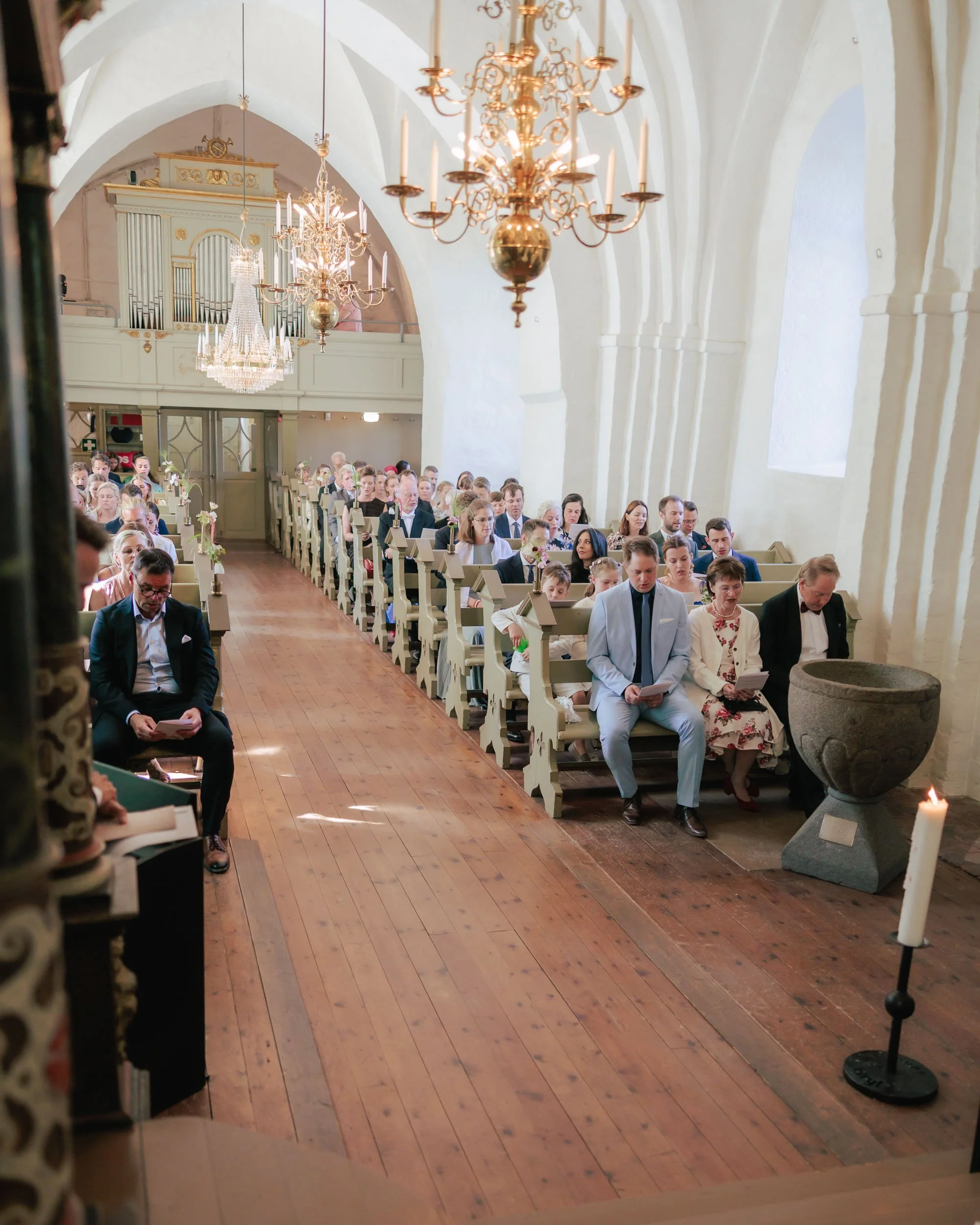 Människor sitter i bänkar i en kyrka under ett bröllop eller liknande ceremoni, med kristallkronor och stora fönster i bakgrunden.