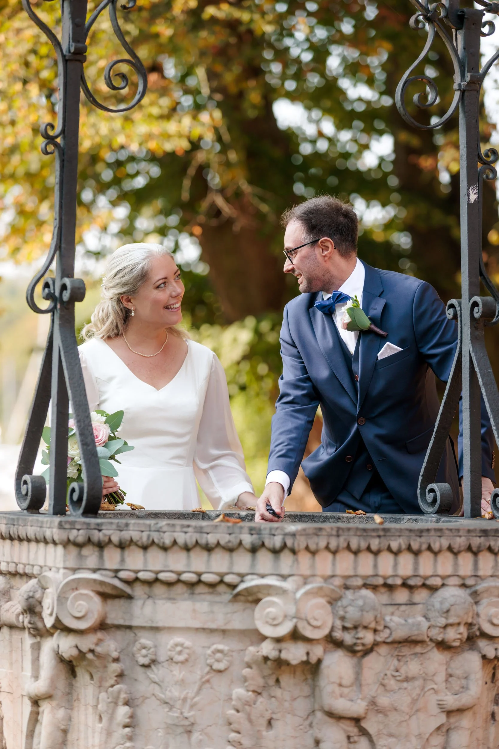 En brud och brudgum ler mot varandra under ett träkors utomhus, omgivna av höstfärgade träd.