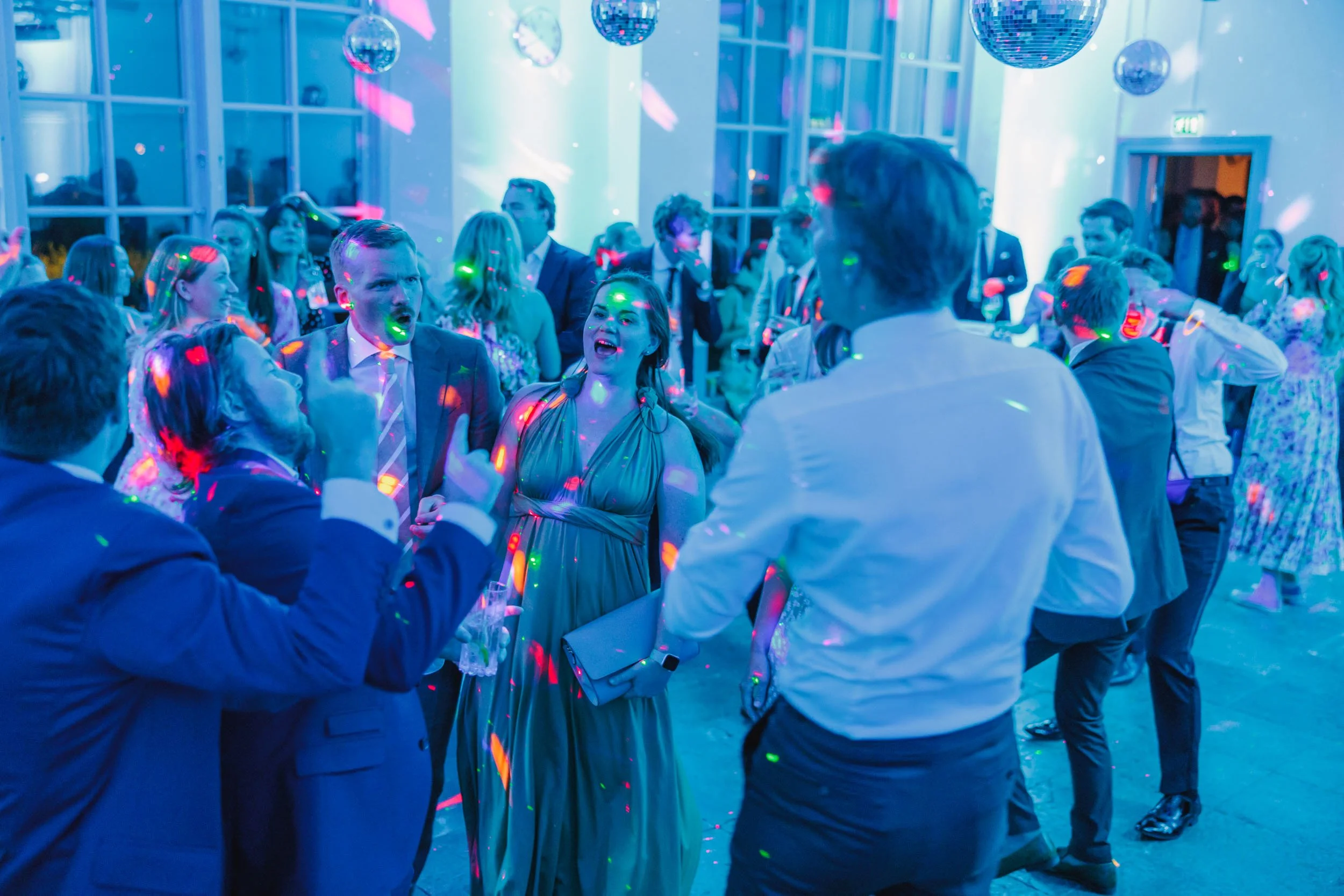 Flera personer som dansar och pratar på ett festligt tillfälle i ett rum med fönster och discokulor, med färgglada ljus och musik.