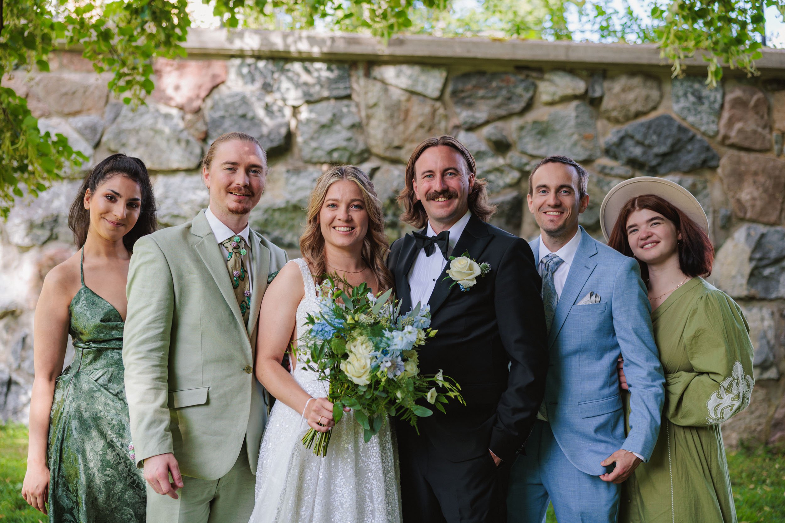 Grupp av sju personer, inklusive ett brudpar, poserar utomhus framför en stenvägg under en festlig tillställning.