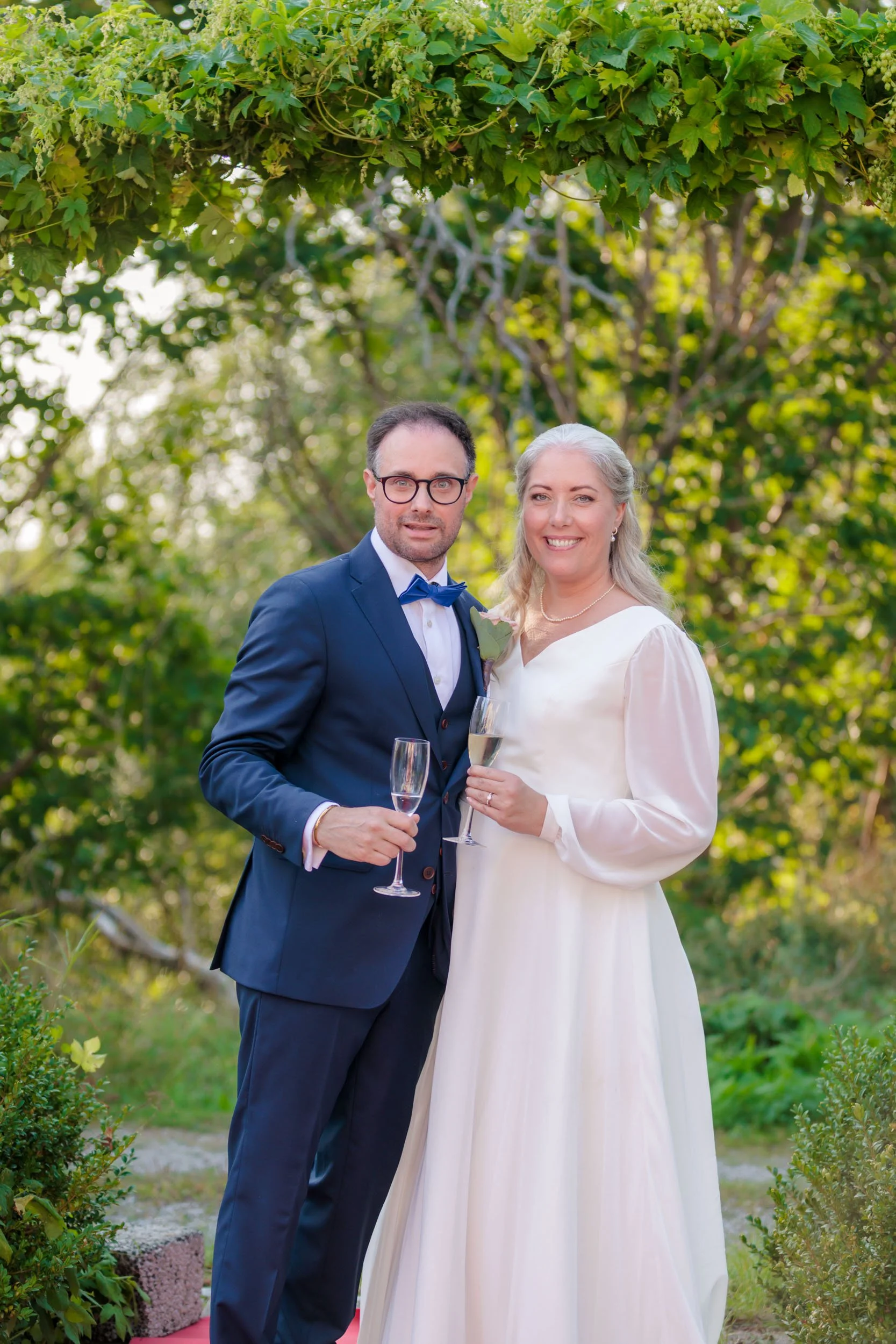 En man och kvinna i bröllopskläder som står utomhus och håller glas med champagne, omgivna av gröna träd och buskar, firar bröllop.