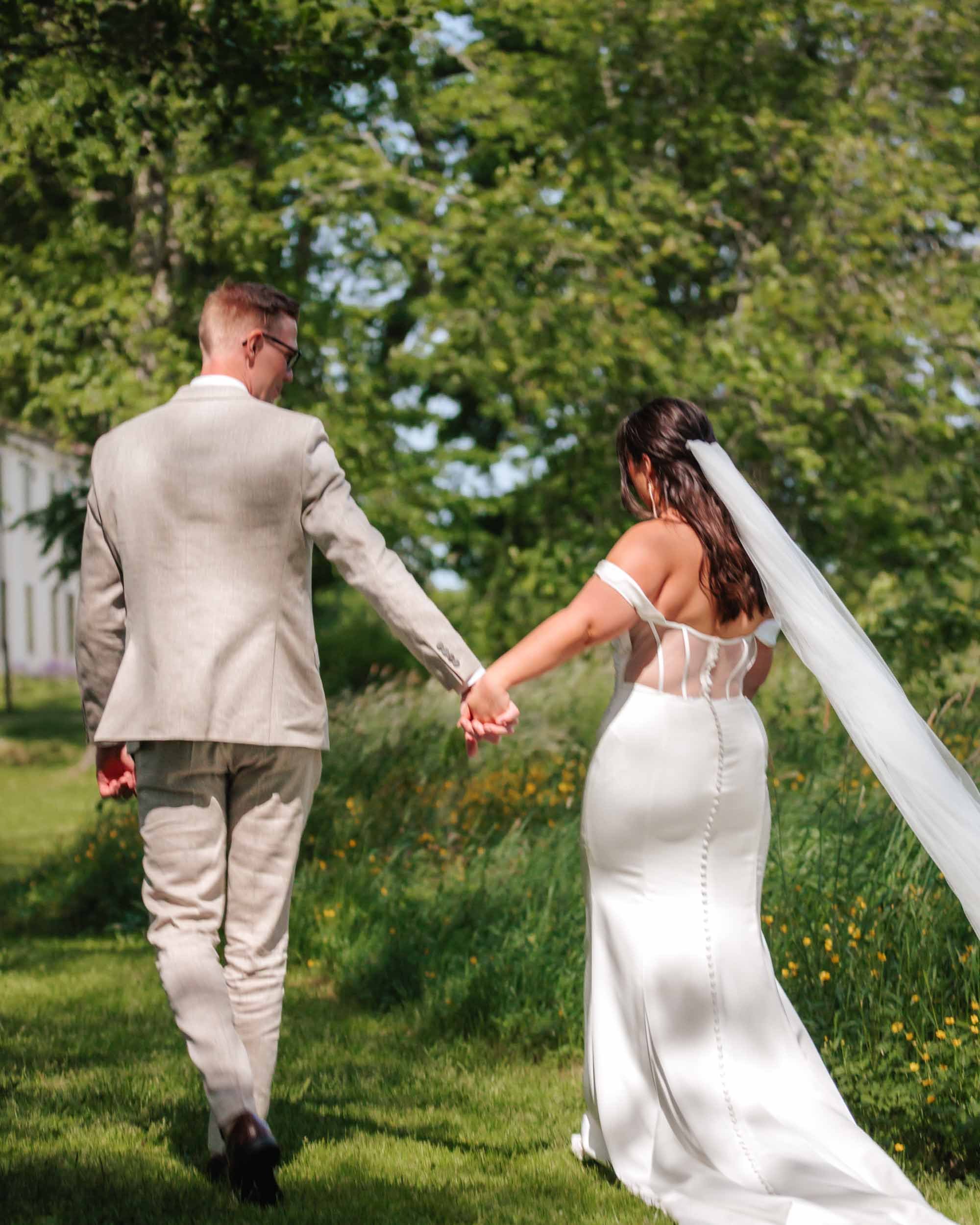 Brud i spetskorsett med slöja och brudgum håller händer bakifrån på promenad i sommargrön miljö