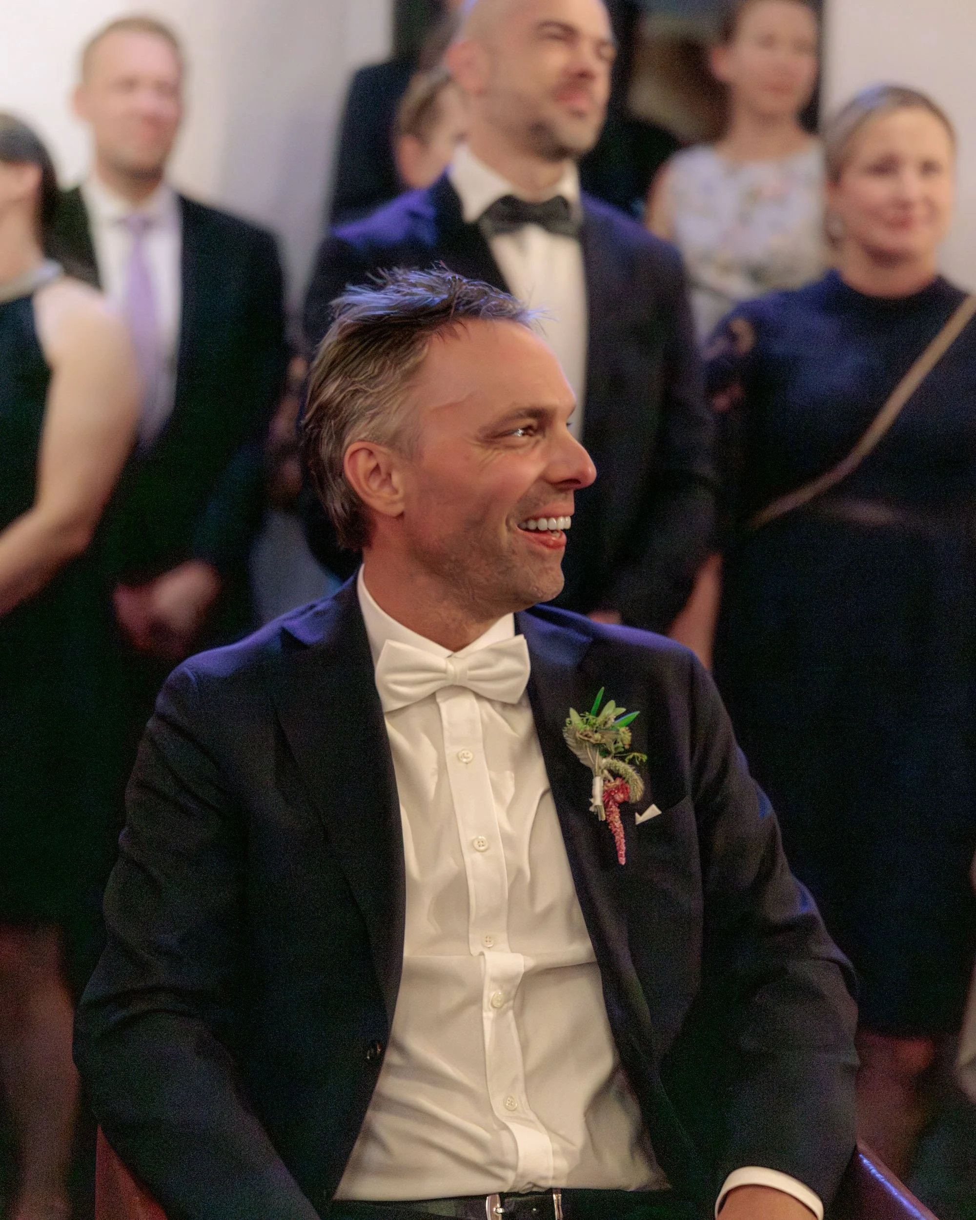 En man i bröllopskläder sitter och ler vid ett bröllop, med flera personer i formella kläder runt omkring honom.