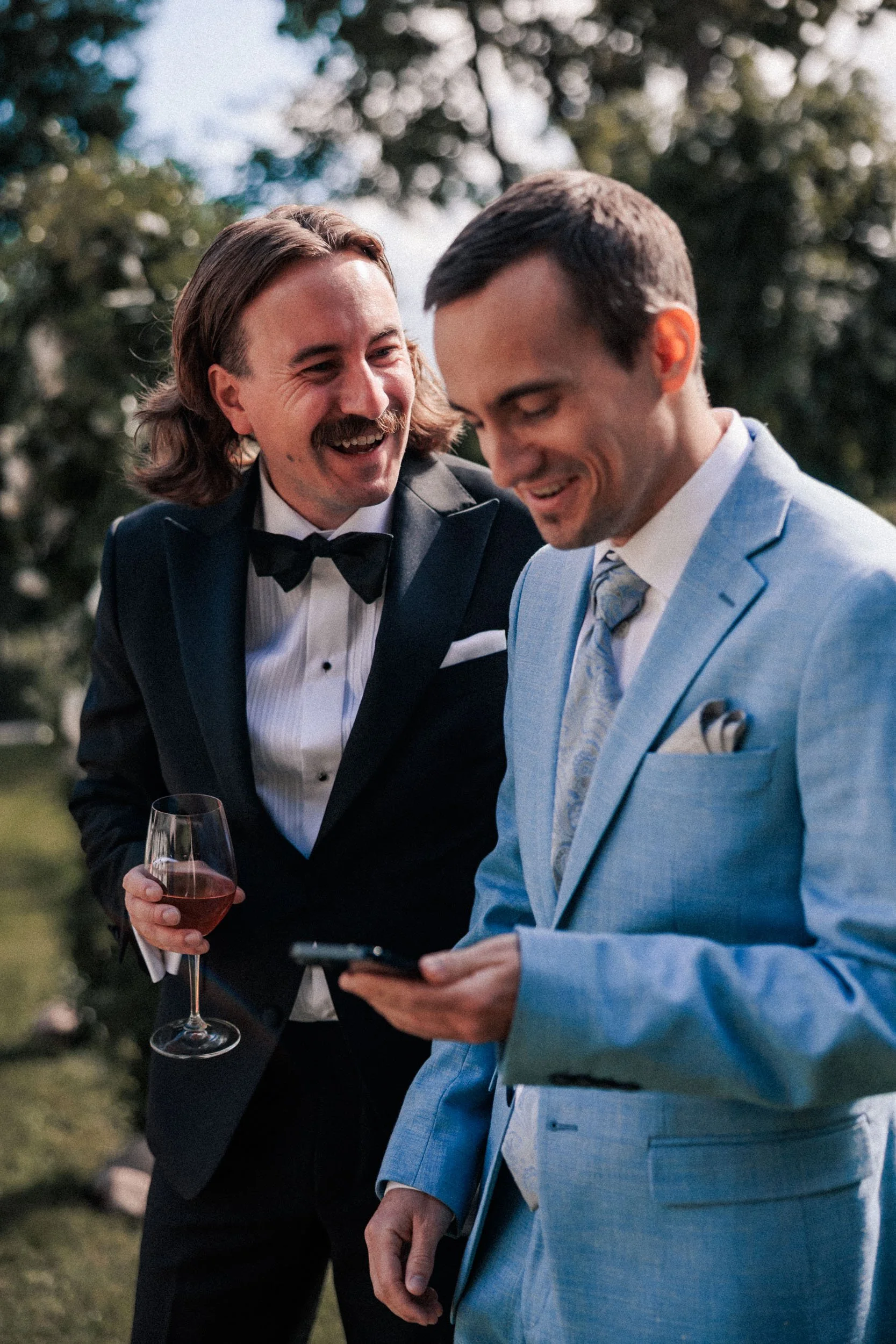 Två män i formella kläder, en i svart smoking och en i ljusblå kostym, ler och tittar på en mobiltelefon under en utomhusfest med grönt träd i bakgrunden.