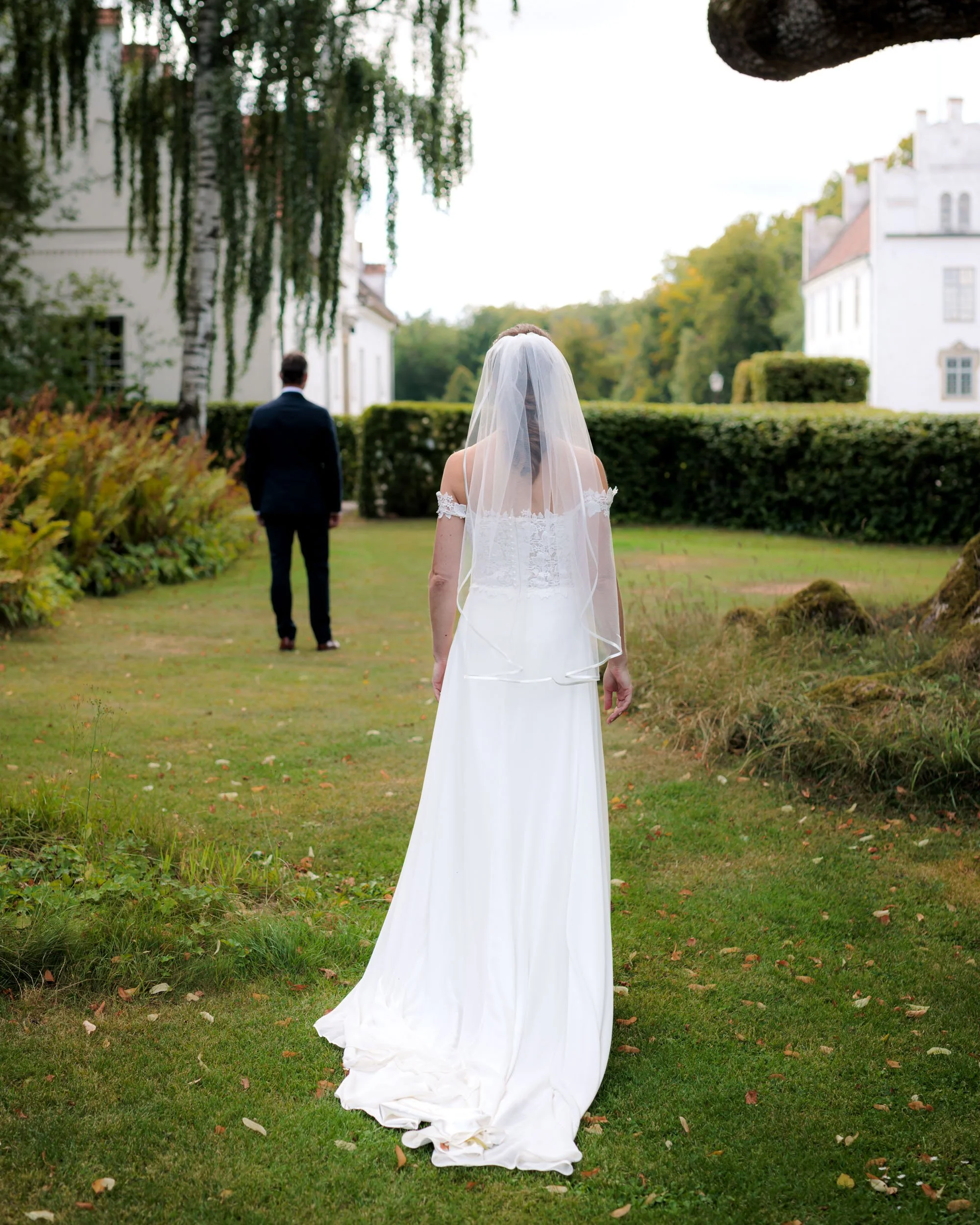 En nygift brud i vit brudklänning och slöja går mot en man i mörk kostym på en gräsmatta utomhus med vita hus och träd i bakgrunden.