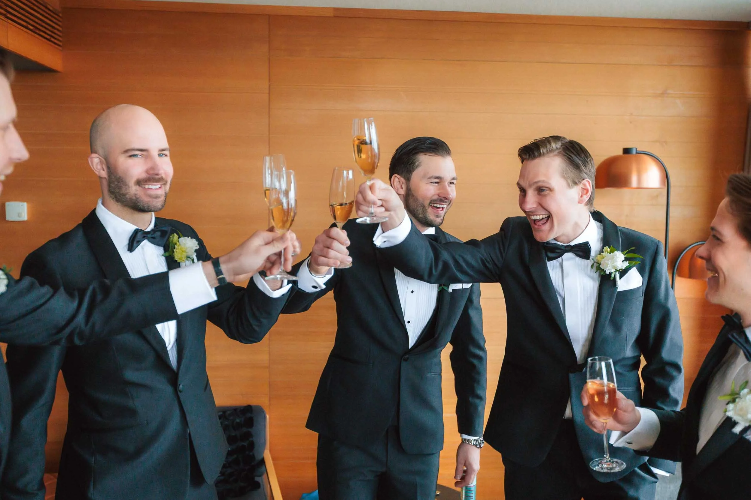 Fem män i formella frackar som firar med skålar av bubbel, skrattar och ler mot kameran i ett trähussesr rum.