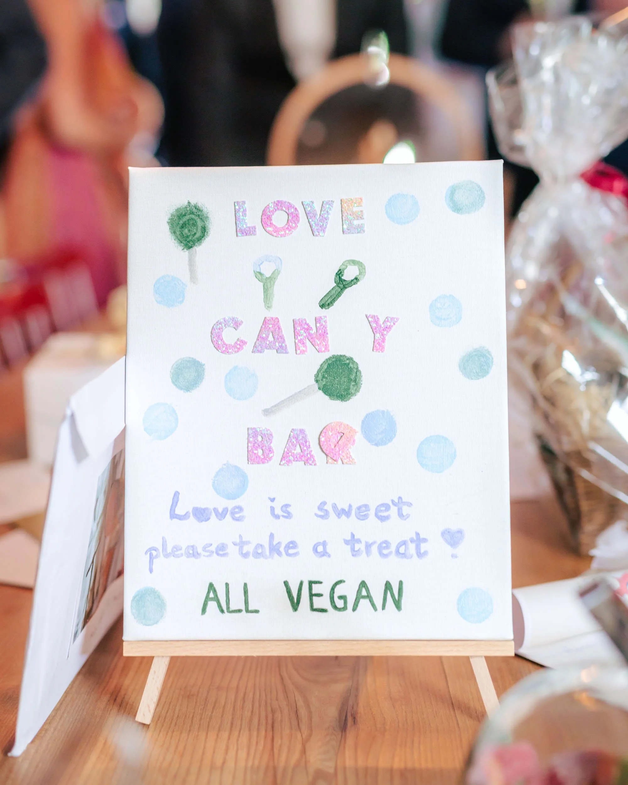 En handskriven skylt med texten 'LOVE CAN Y & BAK Love is sweet please take a treat! ALL VEGAN' på ett bord, omgiven av godis och presenter.