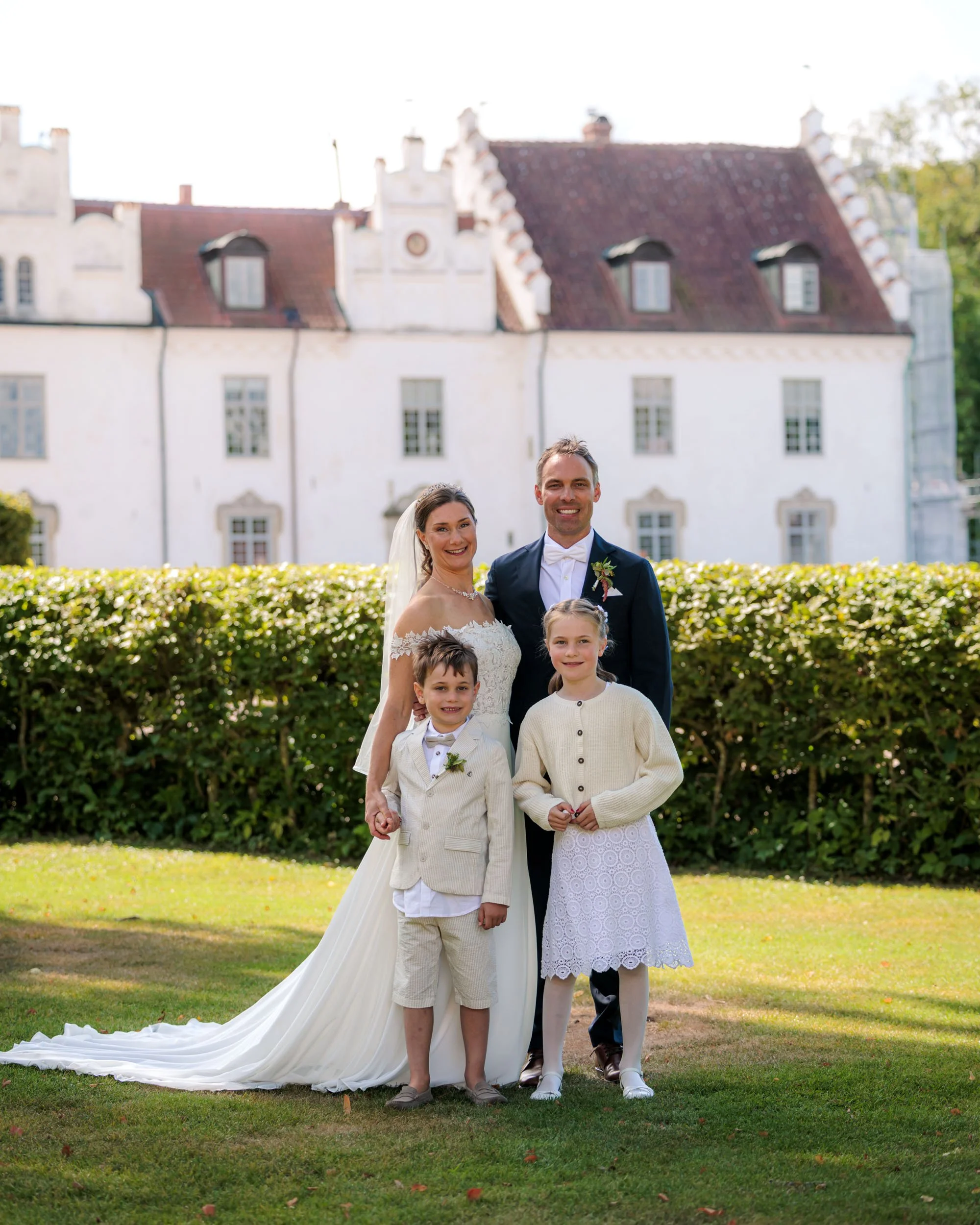 En brud, en gäst och två barn står utomhus på en gräsmatta framför en häck och ett gammalt vitt hus i bakgrunden, alla ler och ser glada ut.