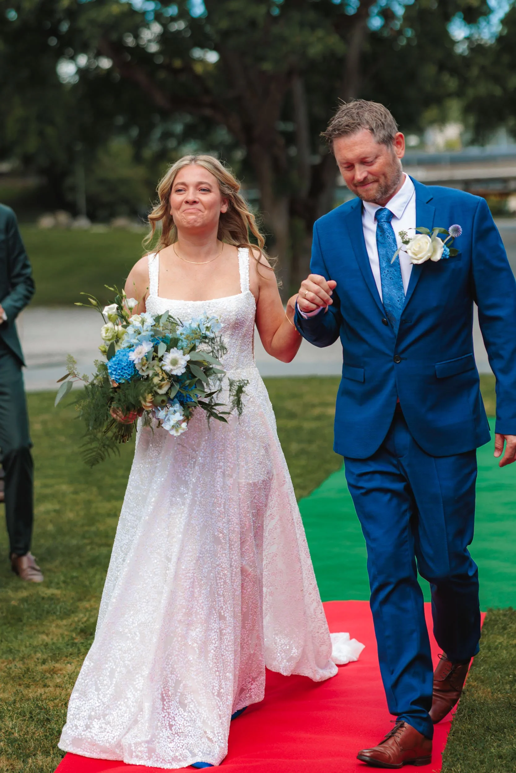 En brud och en man i sinnrika kläder som går på en grön matta utomhus. Bruden håller en stor bukett med vita och blå blommor och de är omgivna av träd och grönt gräs.