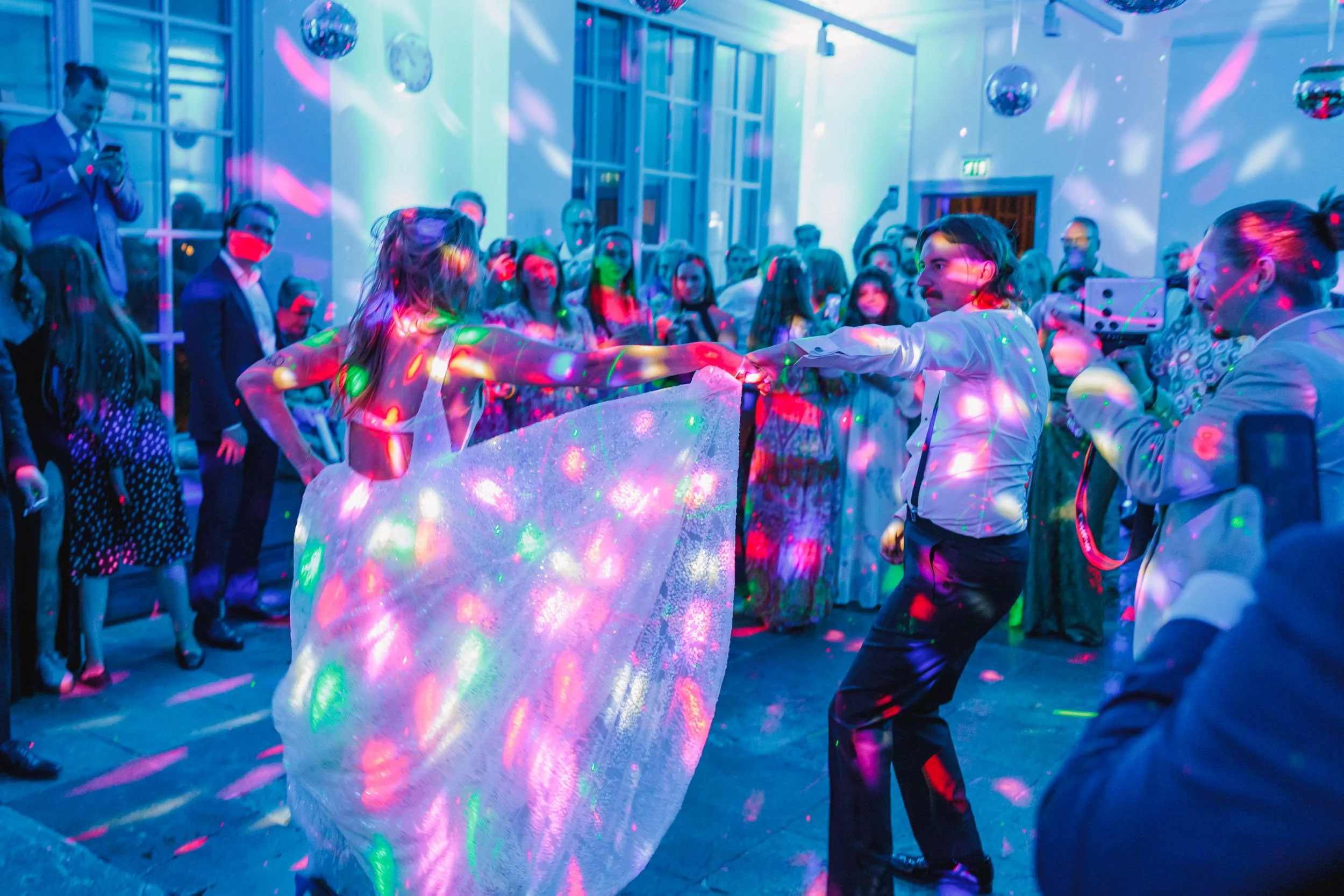 En dansande brud och brudgum i ett rum med färgglad diskobelysning och många gäster.