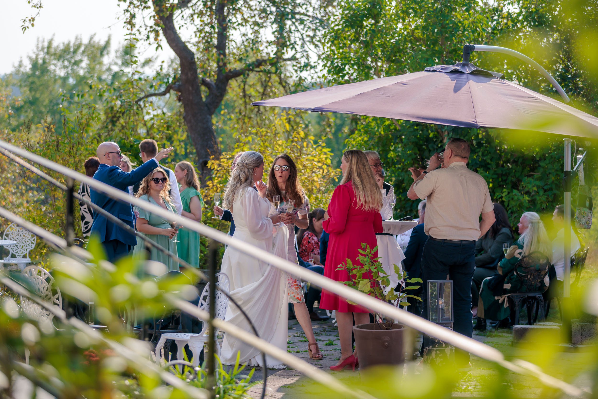 Viggar ett utomhusbröllop där gäster samlas och fikar under ett parasoll, i en trädgård med gröna träd och buskar, på eftermiddagen.