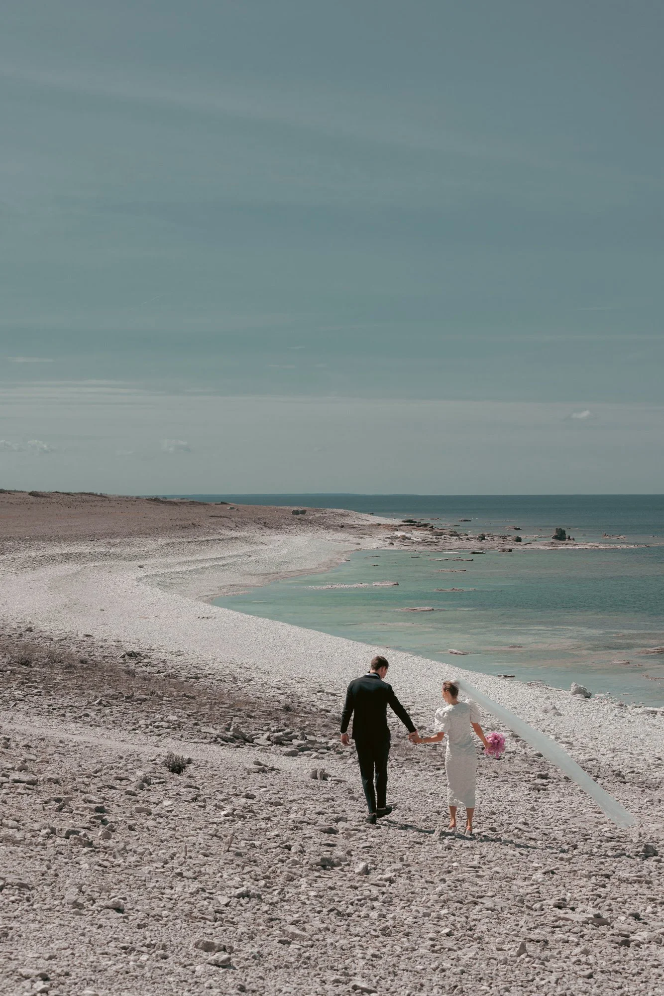 Brudpar promenerar längs stenstrand vid havet under porträttsession på bröllopsdagen