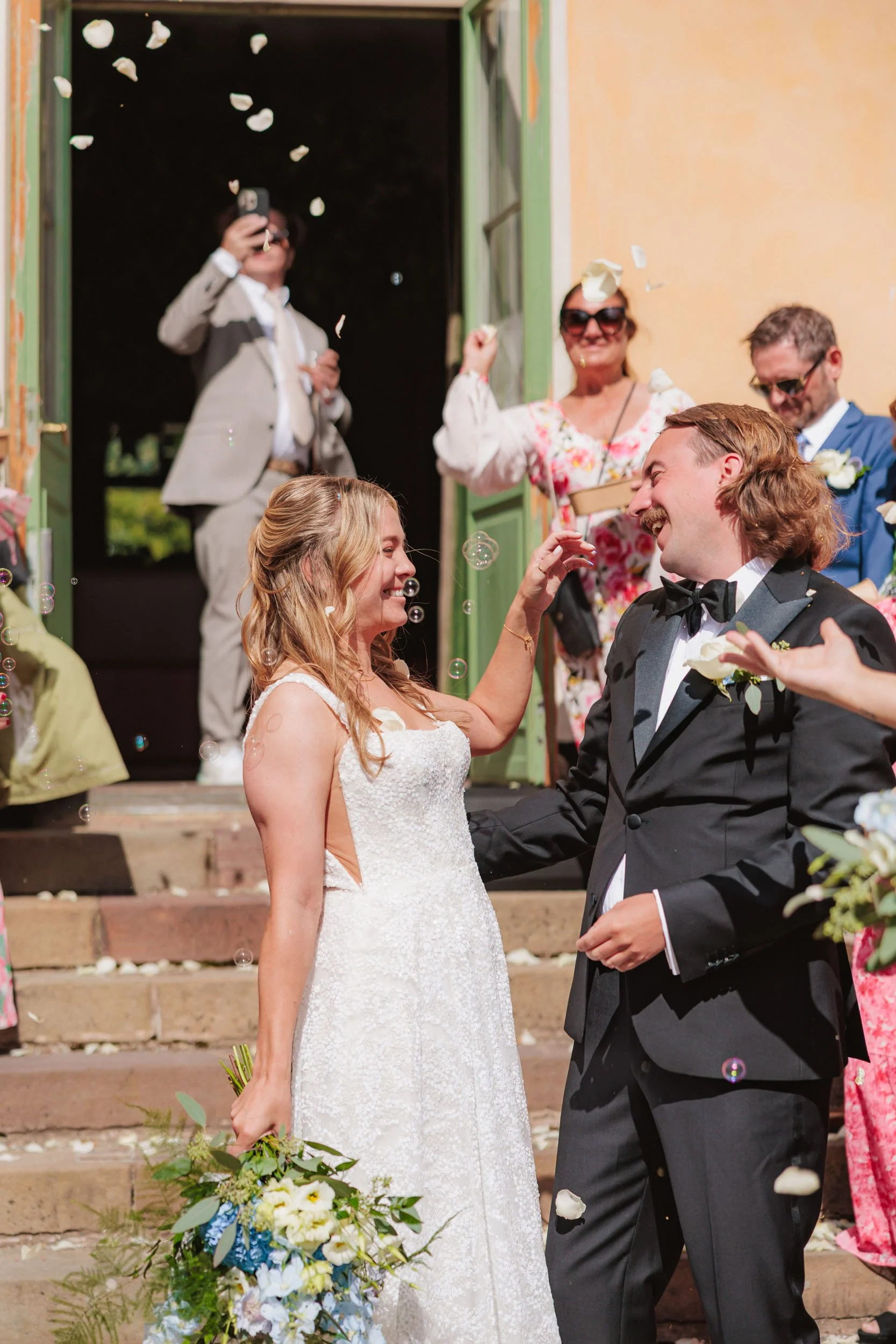 En brud och en brudgum som ler mot varandra under deras bröllopsceremoni, med gäster i bakgrunden som kastar ris och blomblad.