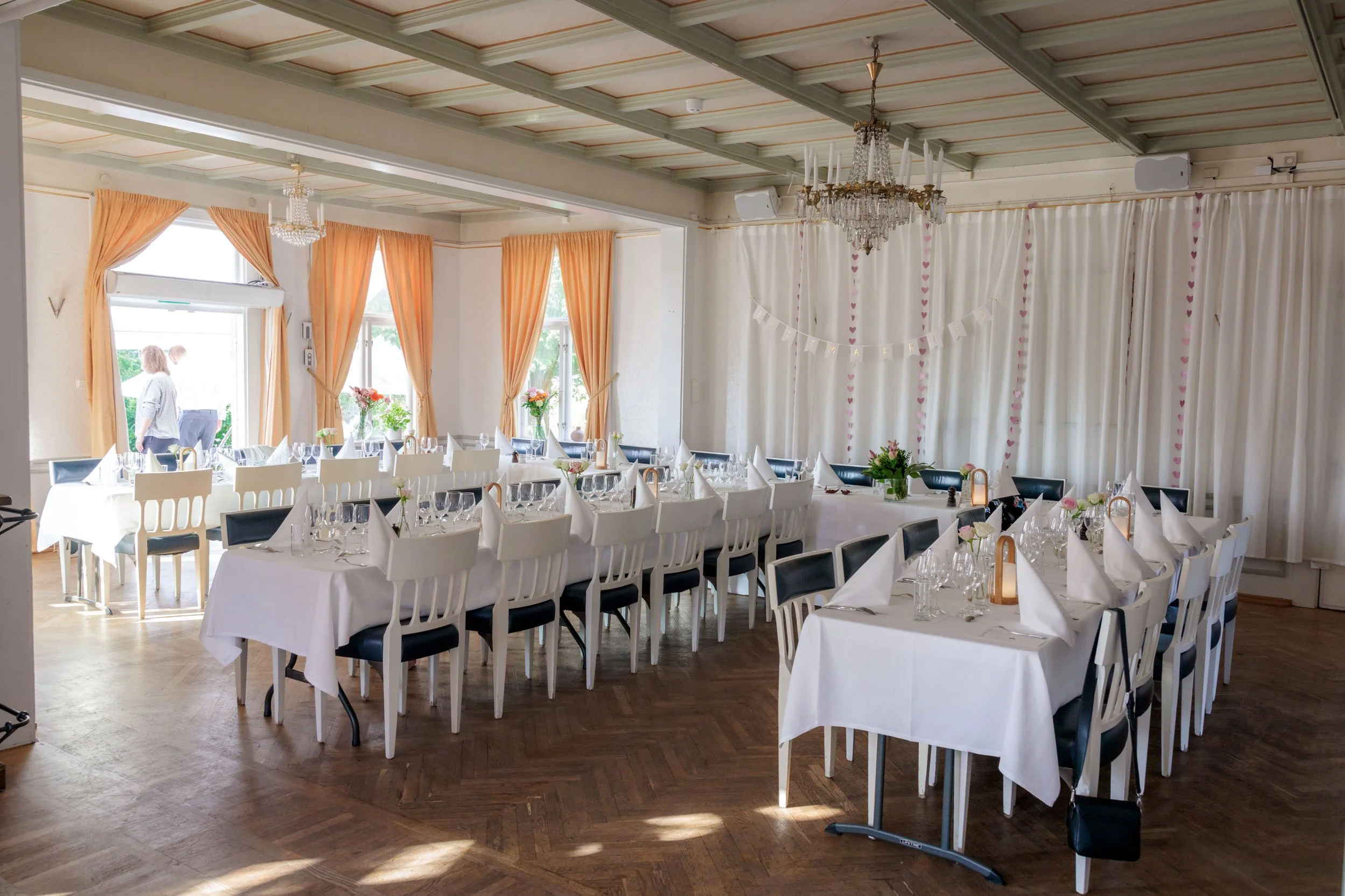 Innredning för ett festligt tillfälle med dukade bord, vita och svarta stolar, blomsterarrangemang, ljuslyktor, och ett draperat bakgrundsskydd med hjärtdekorationer i ett ljust rum.