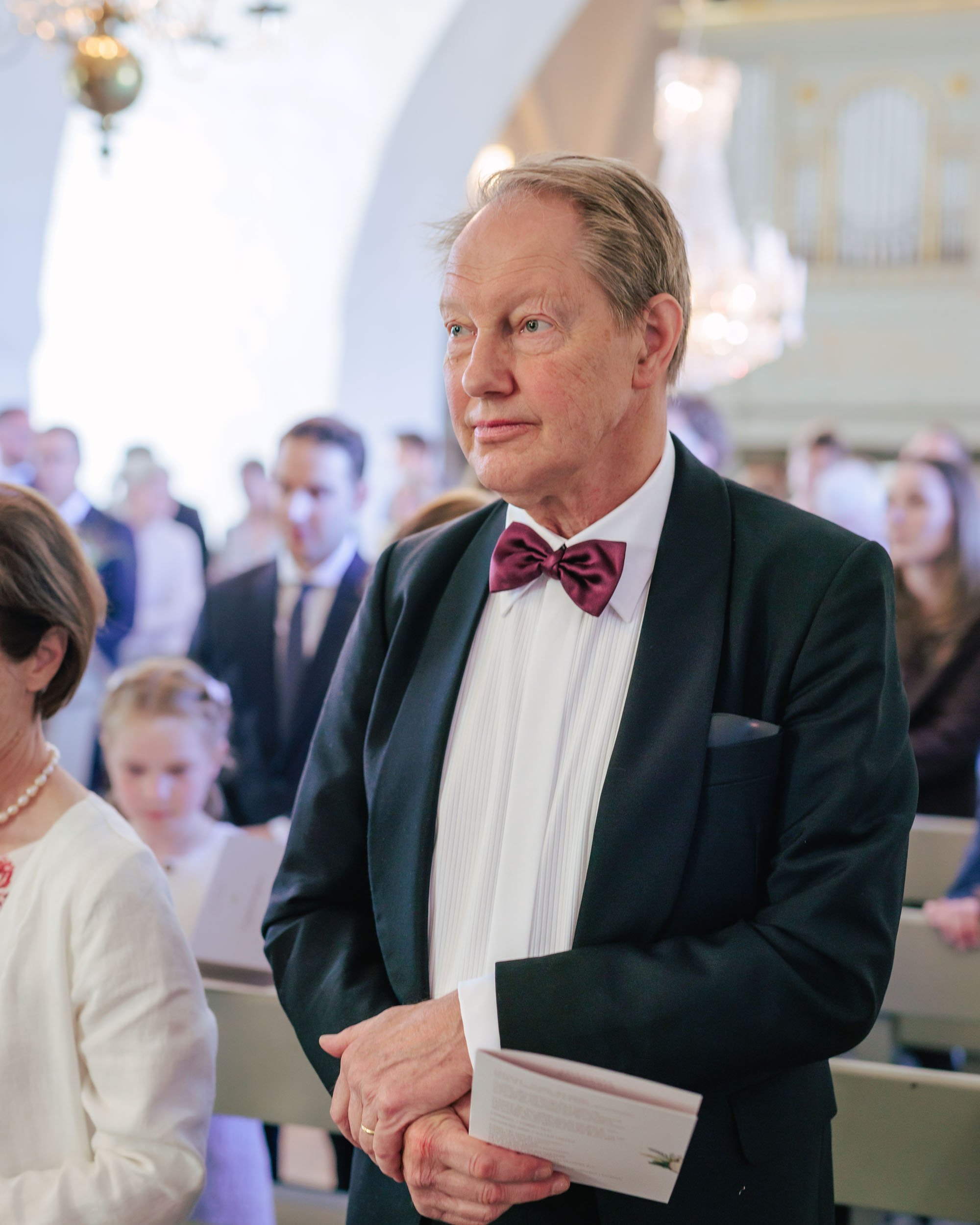 En äldre man i svart tuxedo med vit skjorta och bordeauxfärgad fluga, står i en kyrka under ett bröllop, håller ett informationsblad.