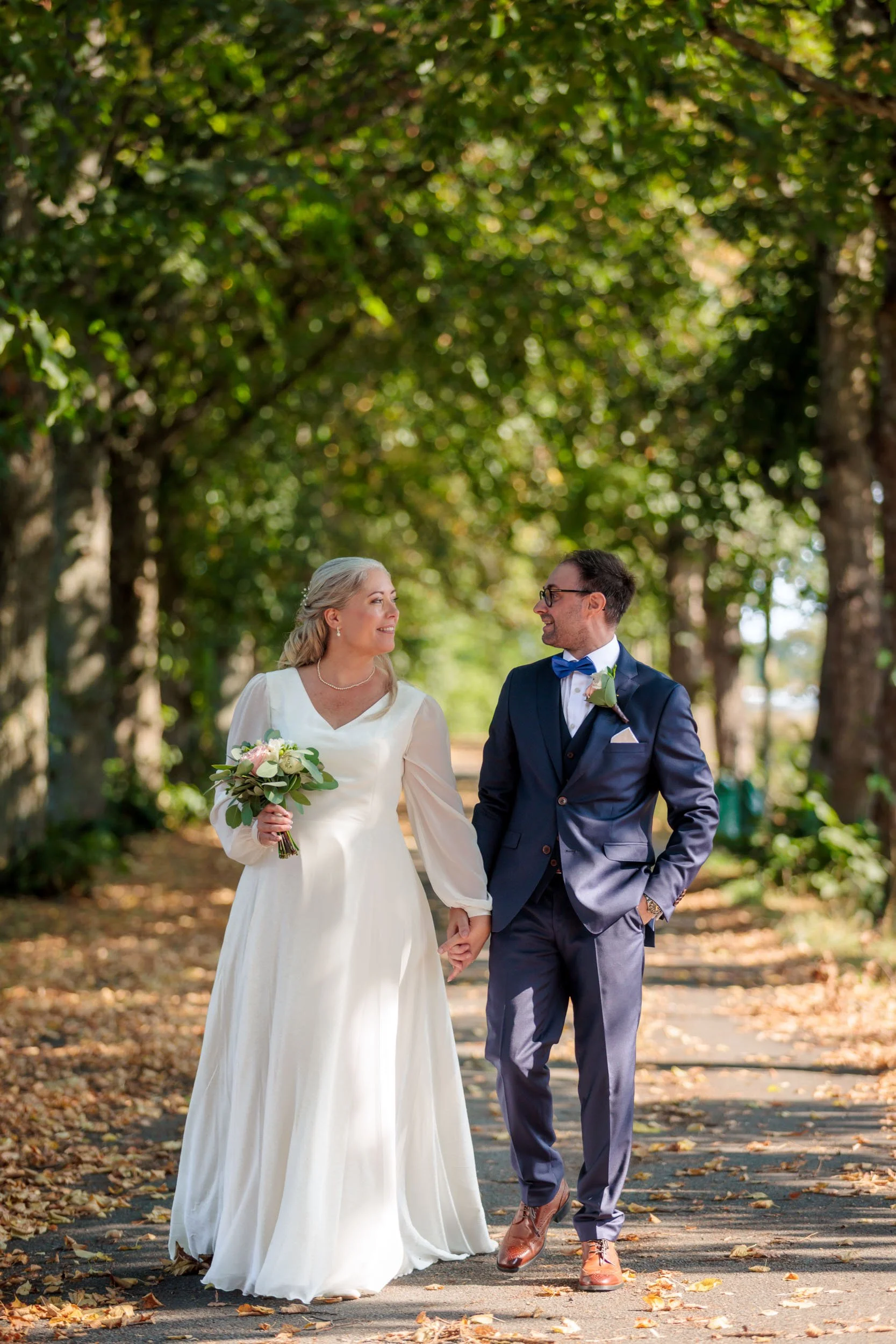 En brud och brudgumma som håller hand och går på en trädallé med höstlöv, ler i bakgrunden, solen skiner.