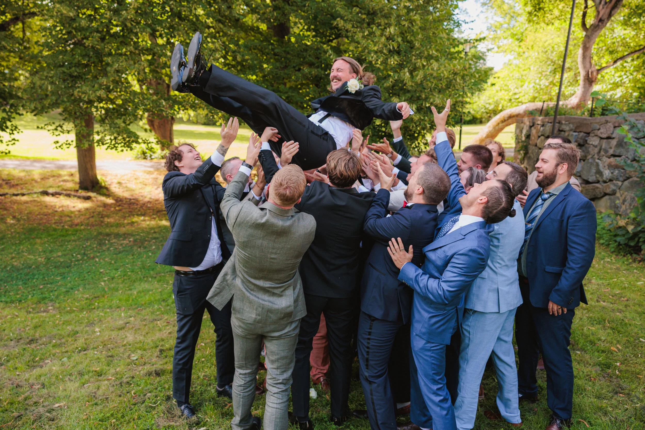 En grupp människor i kostymer som firar en brud i en park, de lyfter upp honom i luften med glädje.