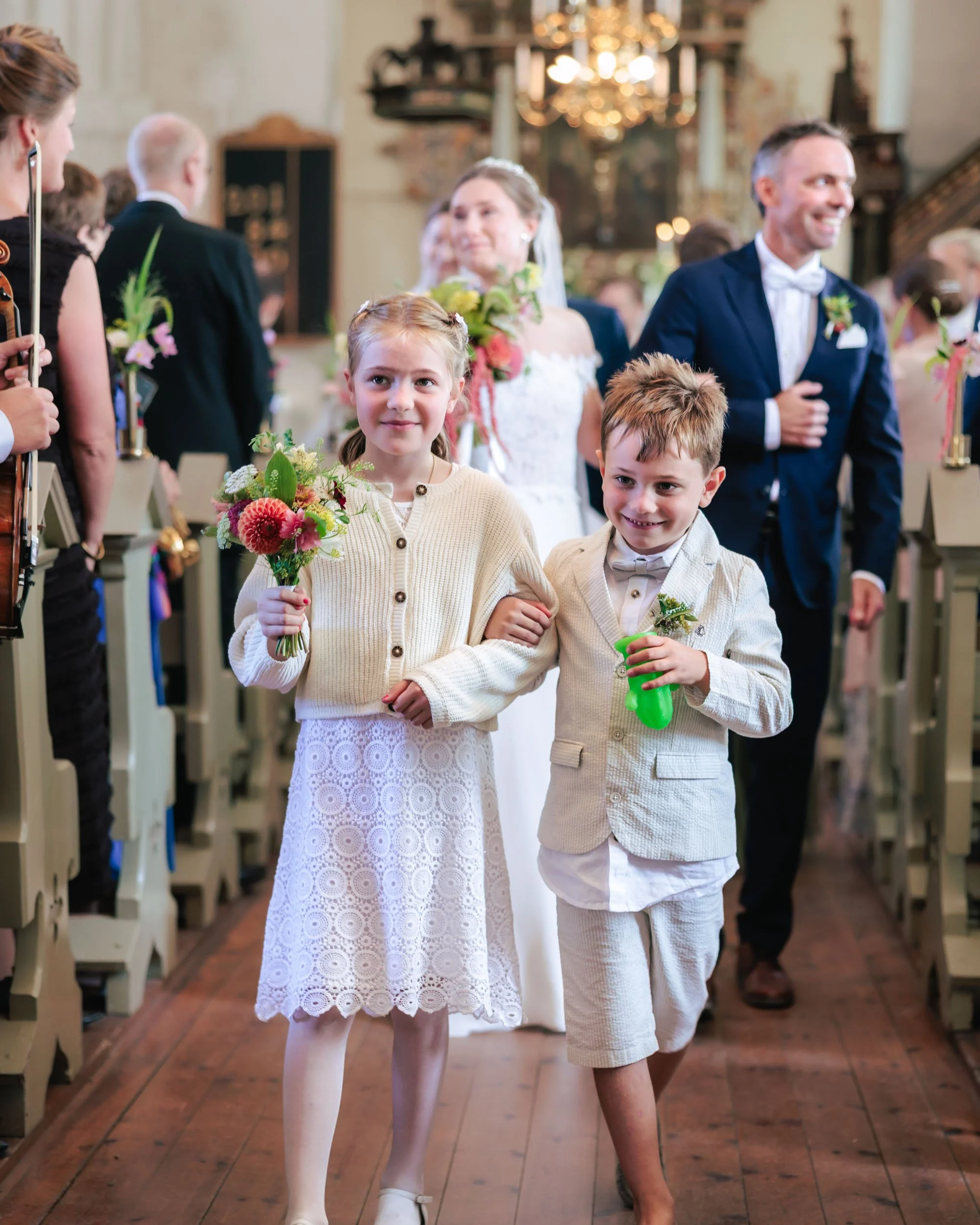 Två barn, en flicka och en pojke, i bröllopskläder som går ner för kyrkgången, omgivna av gäster och ett brudpar i bakgrunden.