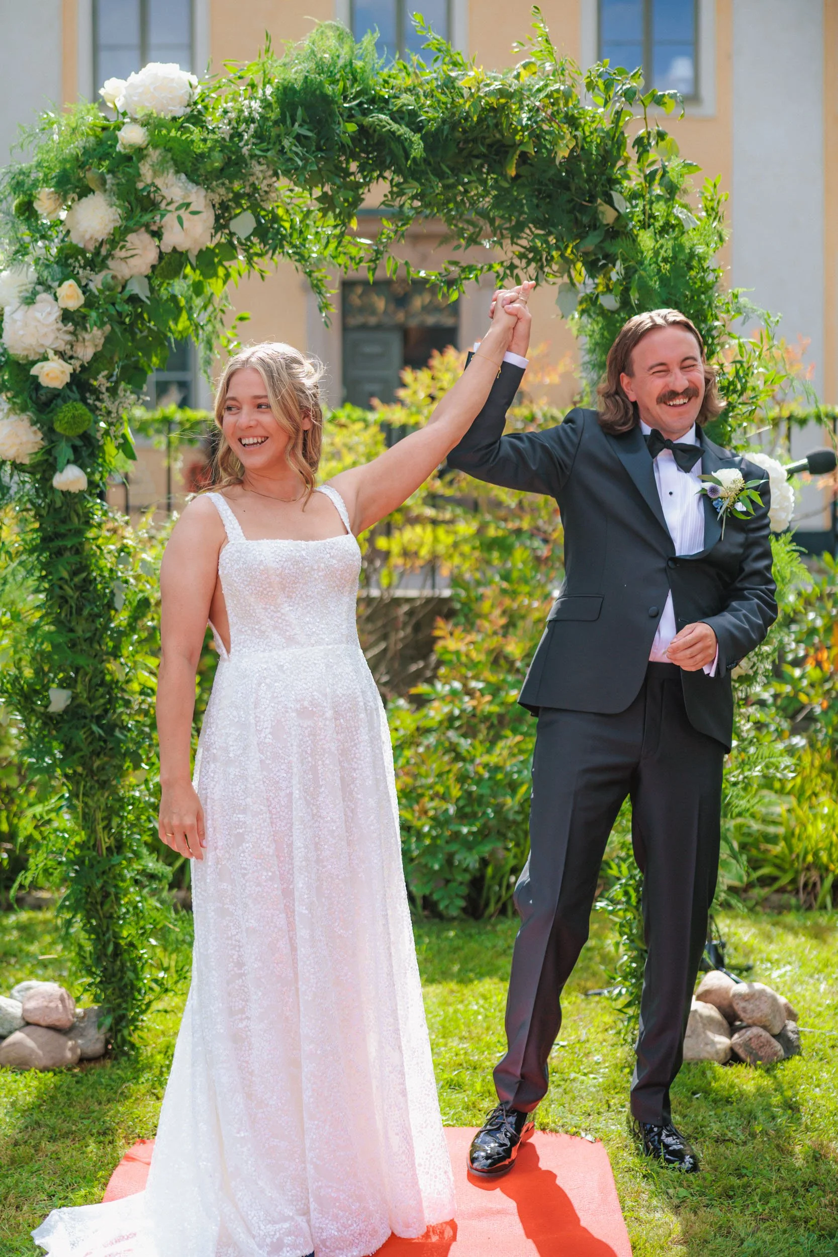 En brud och en bröllopsfotograf som är glada och håller handen uppe i ett bröllop, utomhus vid ett grönt lövverk och en blomsterdekoration i bakgrunden.