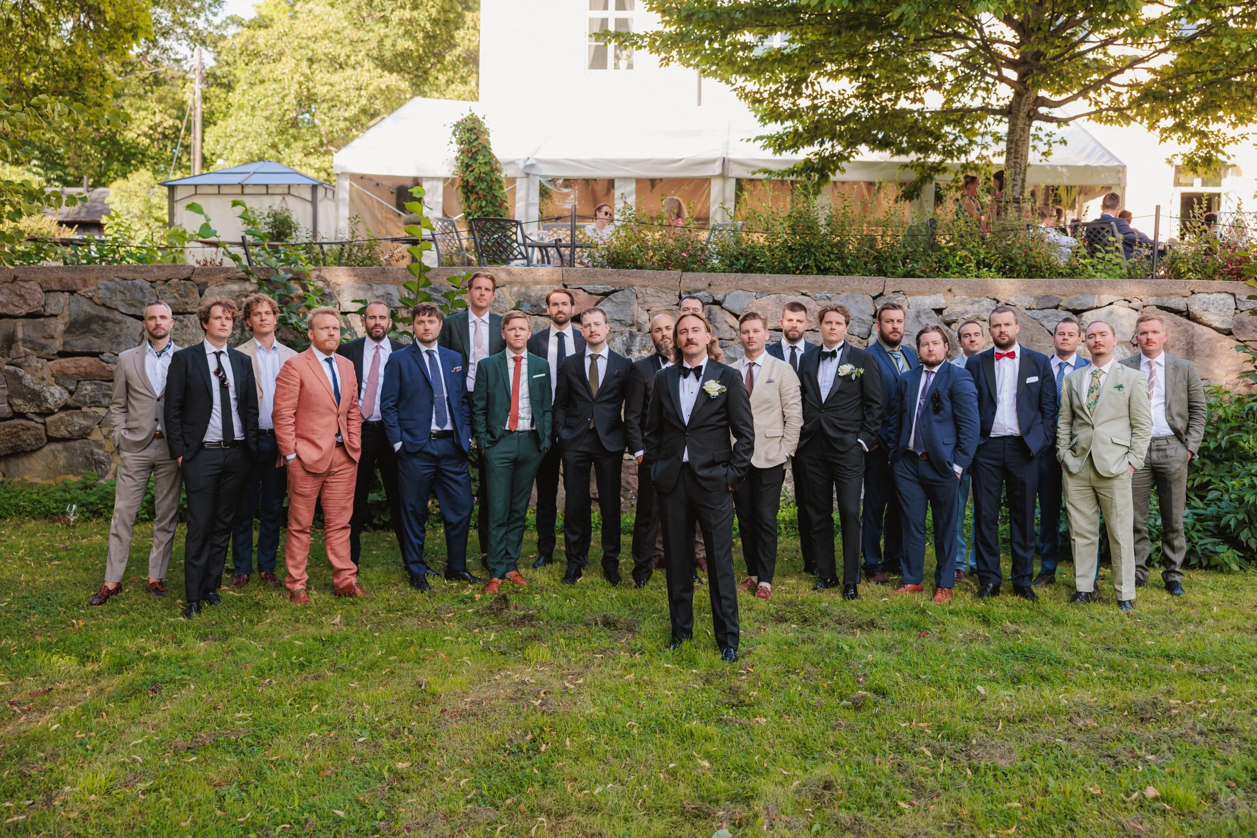 Gruppbild av män i formella kläder på gräsmatta vid utomhusbröllop, med en stenmur och ett tält i bakgrunden.