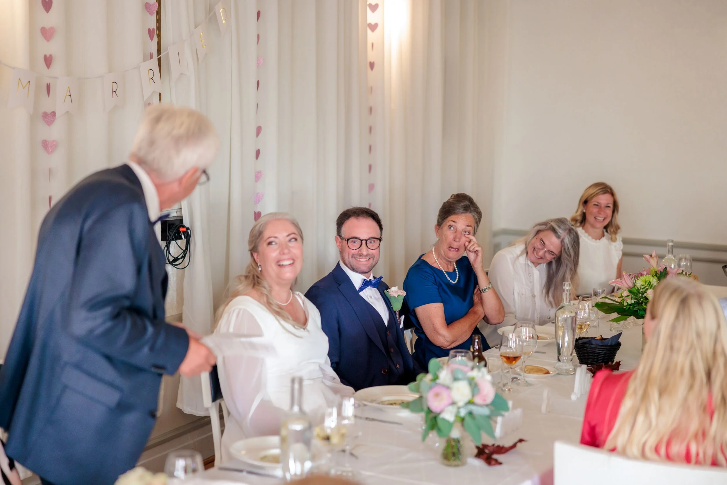 Människor sitter vid ett bord under ett bröllop, med en man som ger ett tal. Bakgrund med hjärtfestade dekorationer och ett band med texten Marry Me.