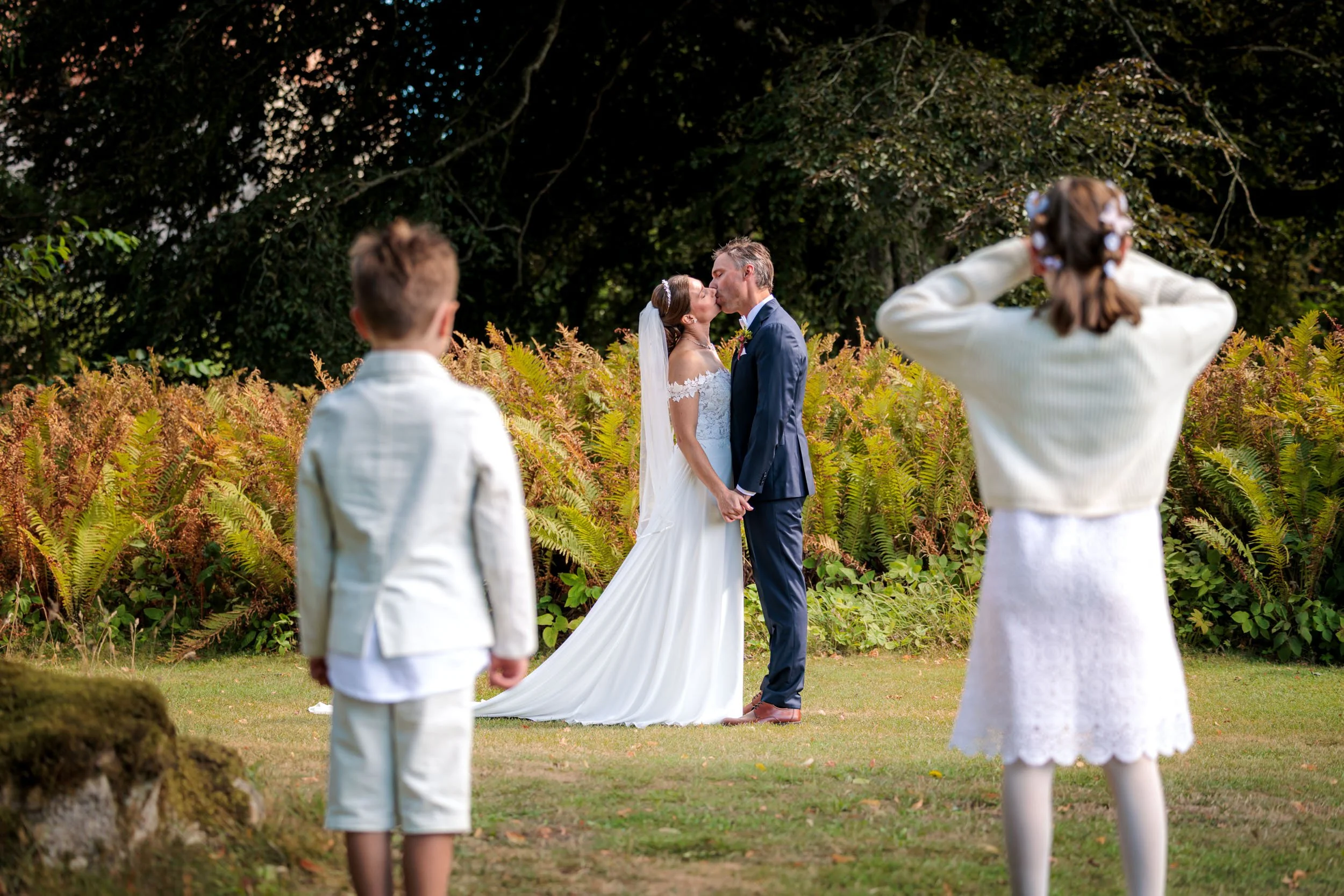 En brud och en brudgum kysser varandra under en utomhusvigning, omgiven av två barn, en pojke och en flicka, på en gräsmatta med gröna växter och träd i bakgrunden.