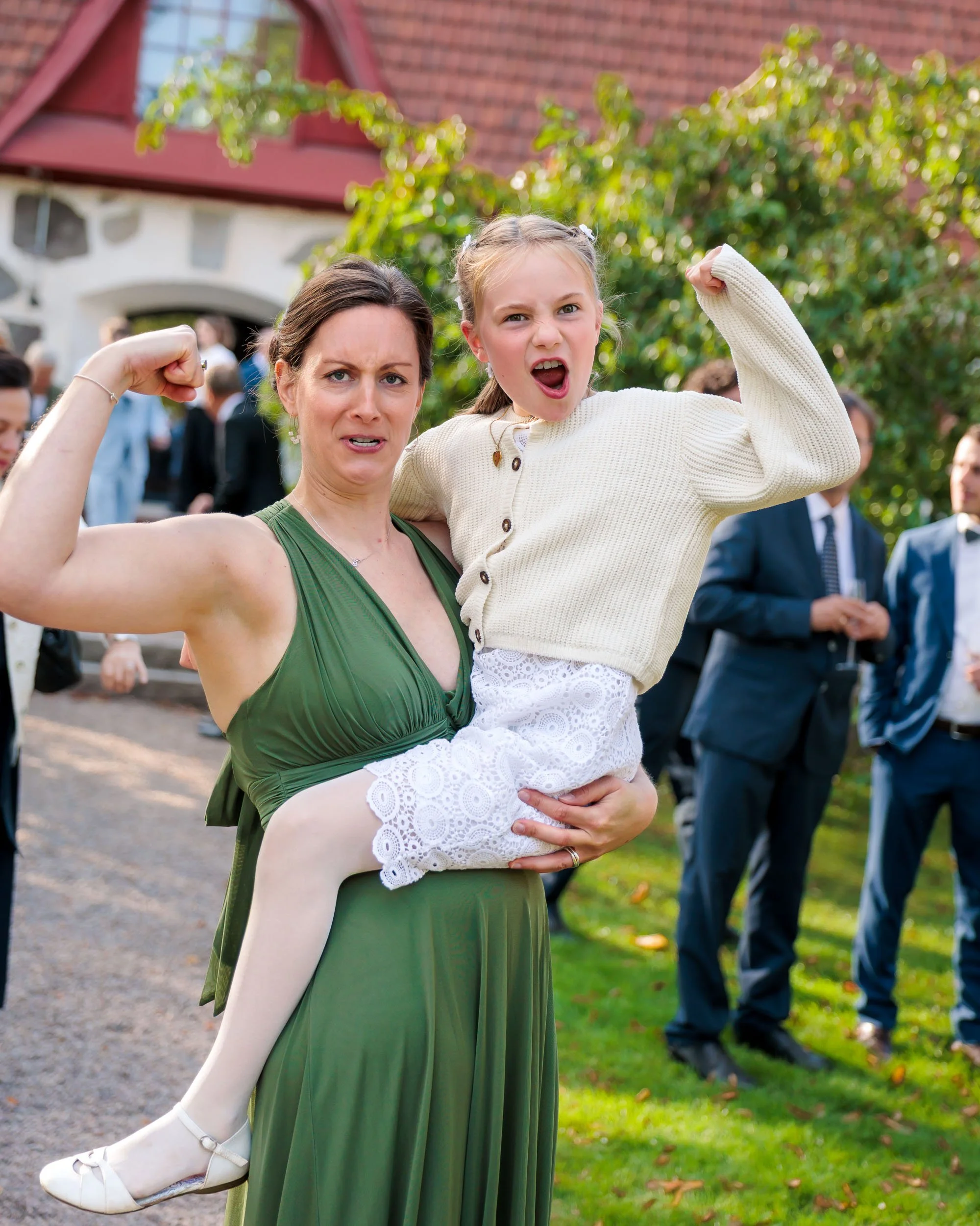 En kvinna i grönt klädd, som håller i en ung flicka, båda gör muskelbice-gest. Bakgrund med andra människor i kostymer och ett hus, utomhus på en gräsmatta.