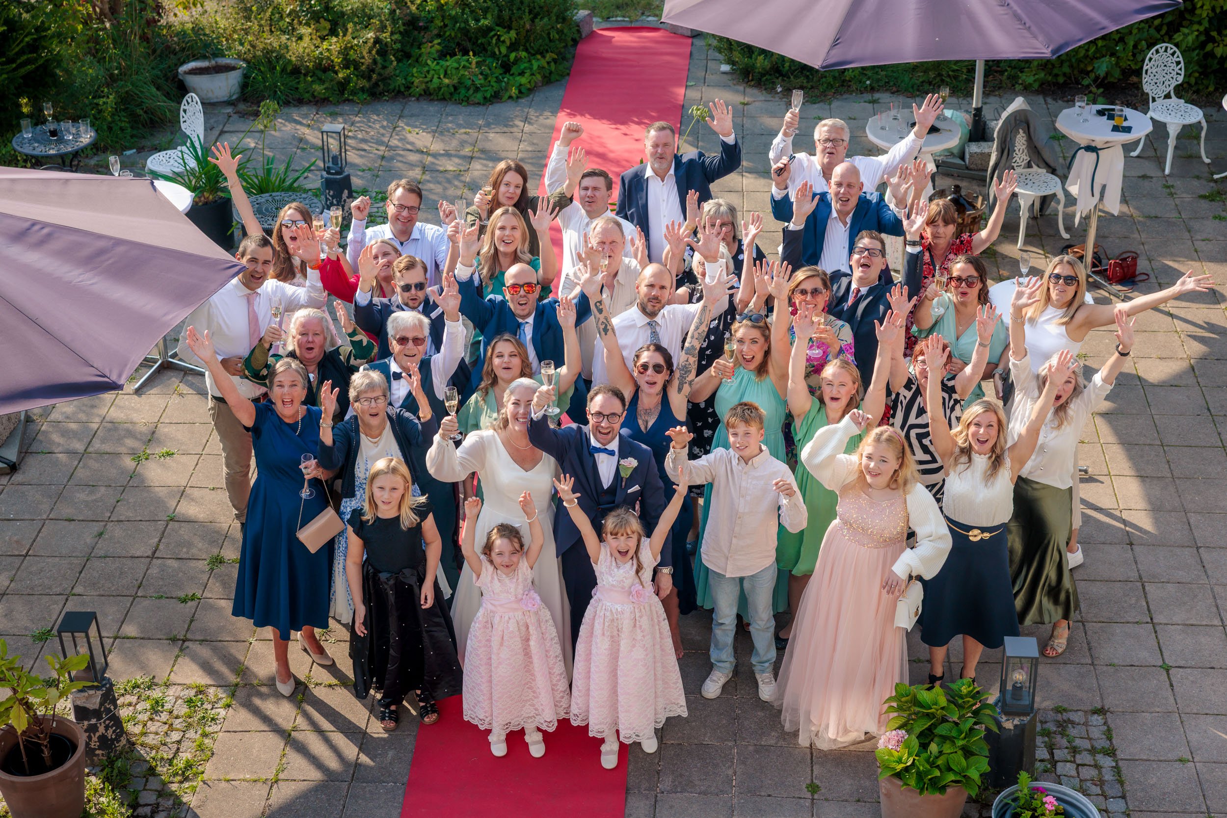 En stor grupp människor som firar på en uteservering, många med glas i handen, skrattar och vinkar mot kameran. De ser glada och festklädda ut, och några barn står framför gruppen.