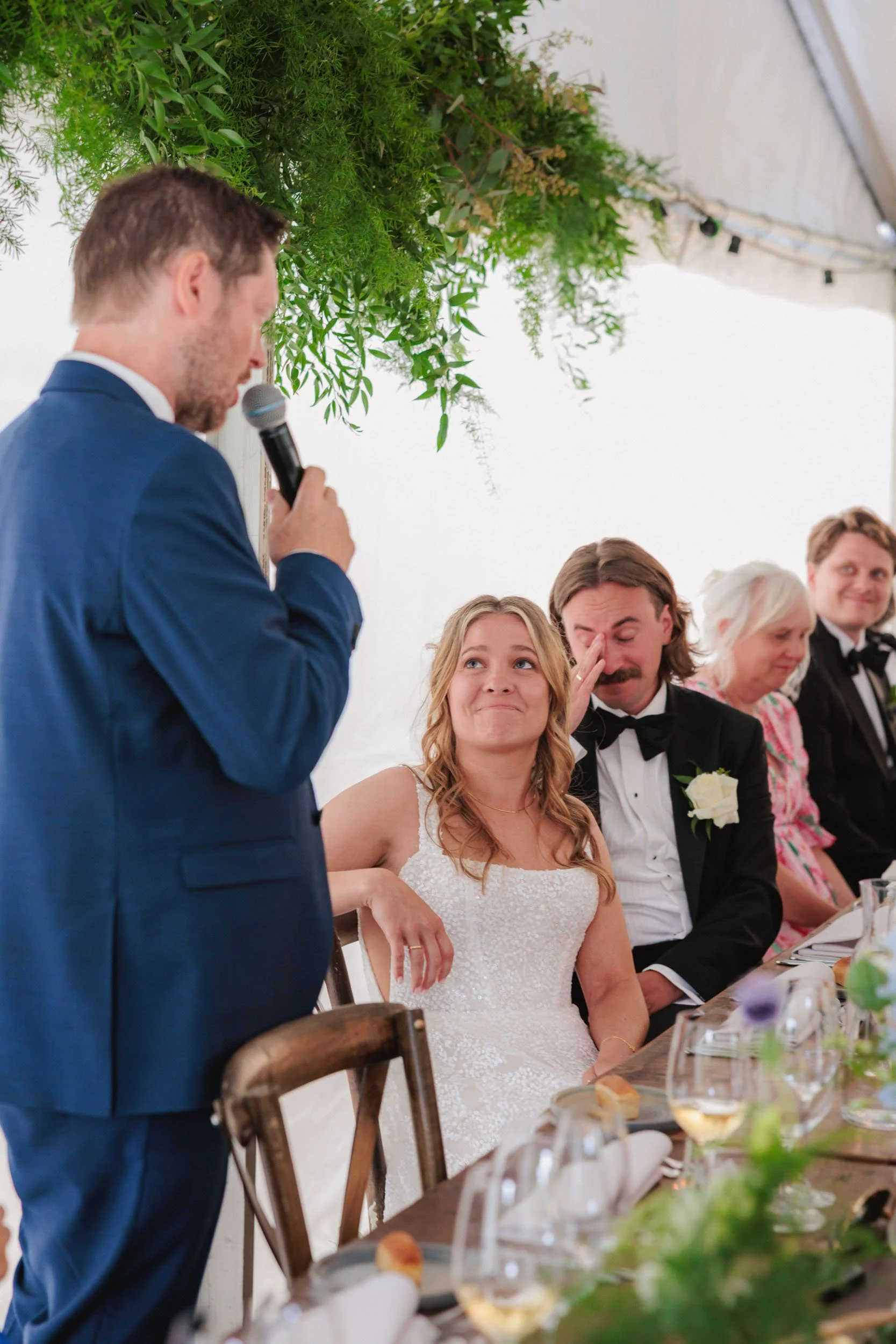 En man i blå kostym håller en mikrofon och talar till ett brudpar vid ett bröllop, där bruden ser rörd ut och gesterar med handen. Brudgummen rör sig till tårar, och flera personer i formella kläder sitter vid ett bord.