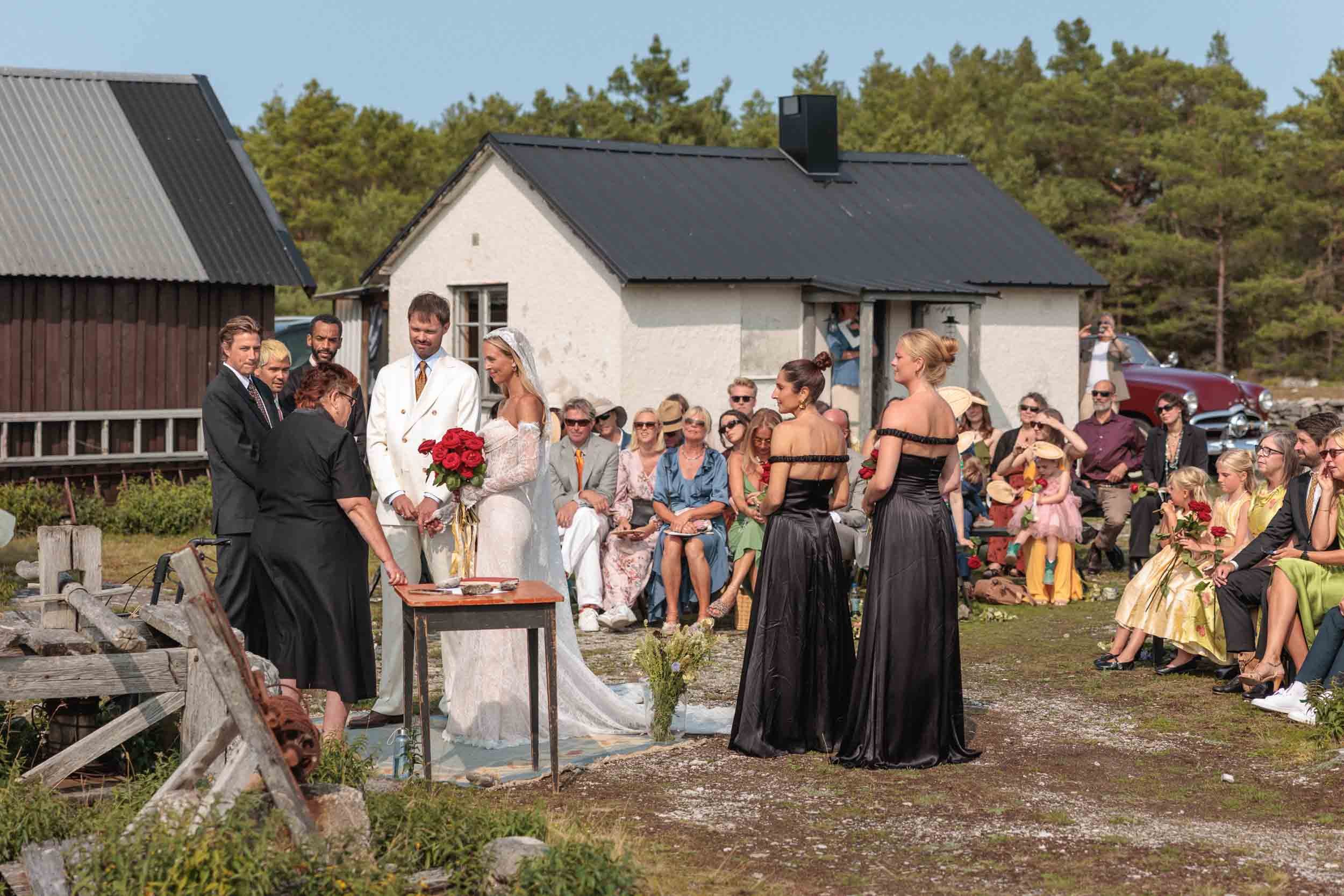 Brudpar vid vigselbordet under utomhusceremoni i lantlig miljö med vita stugor och tallskog