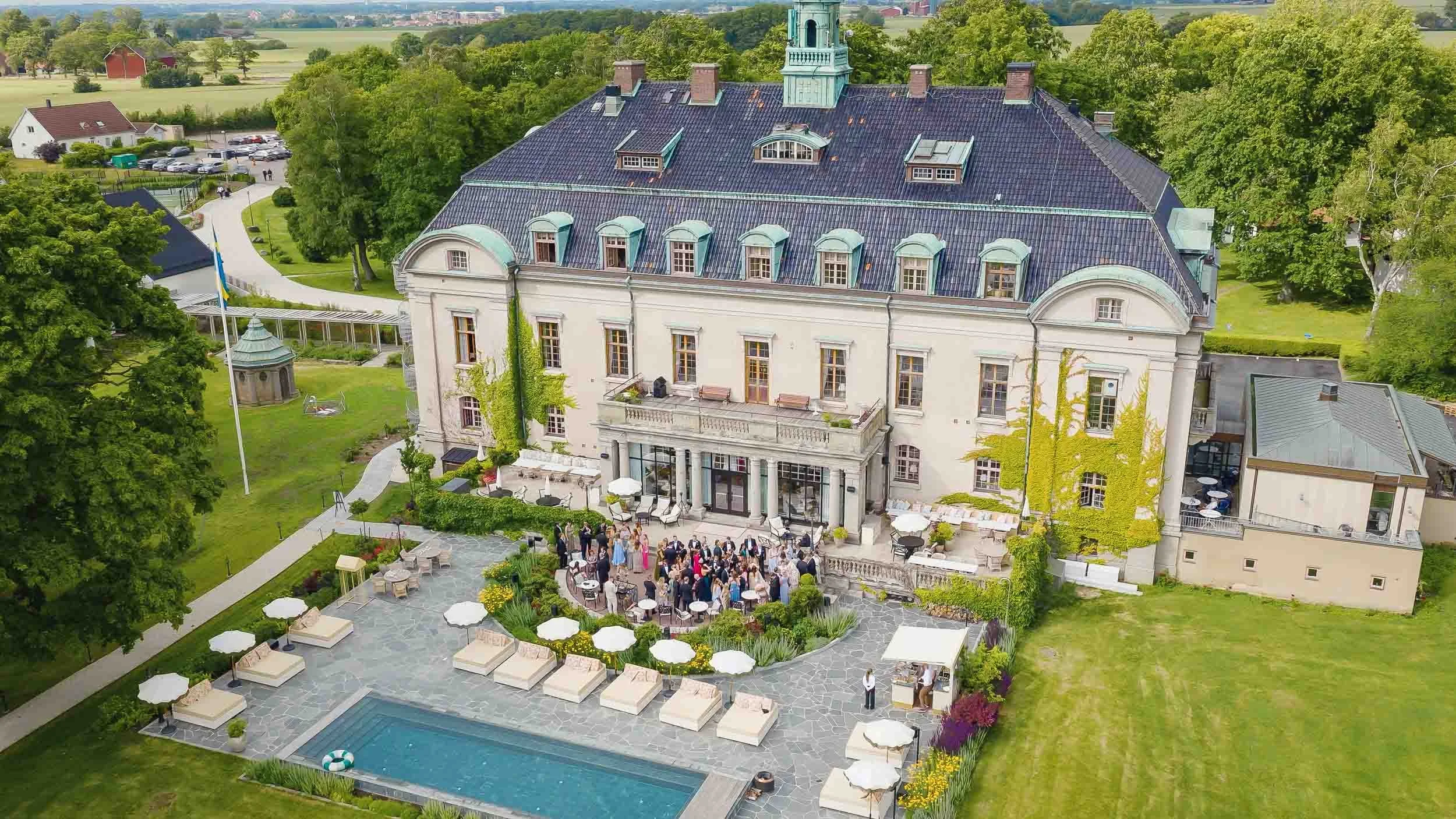 A large, elegant mansion with a garden and an outdoor pool. People are gathering for an event on the terrace in front of the house.
