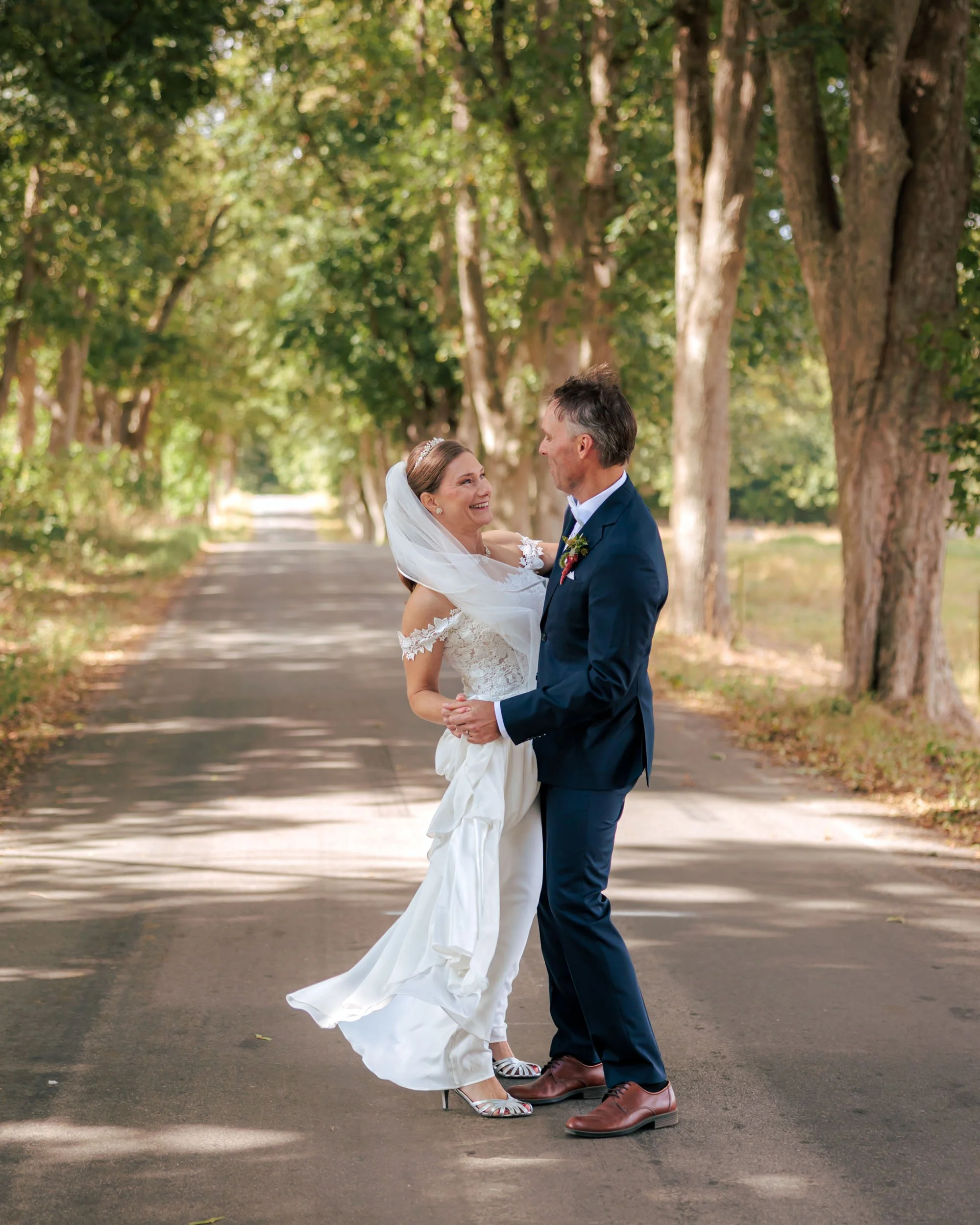 Brudparet står på en trädkantad väg, ler och håller varandra i handen i en utomhusmiljö under dagen.
