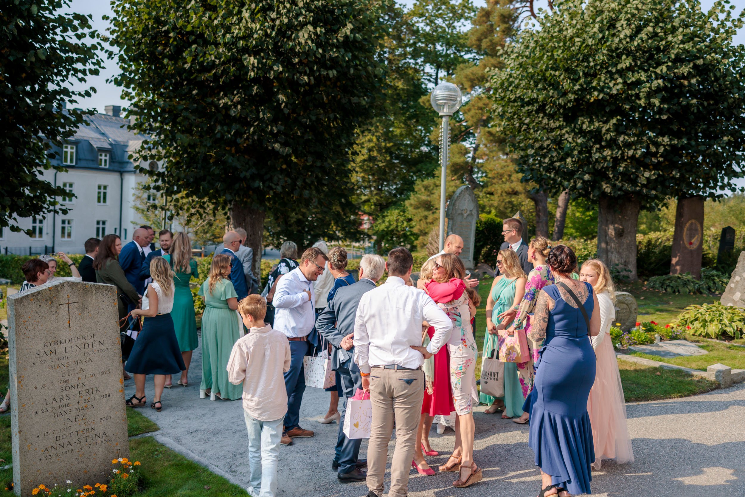 Människor samlade utomhus i en park, många i sommarkläder, några kommunicerar och andra poserar framför gravstenar.