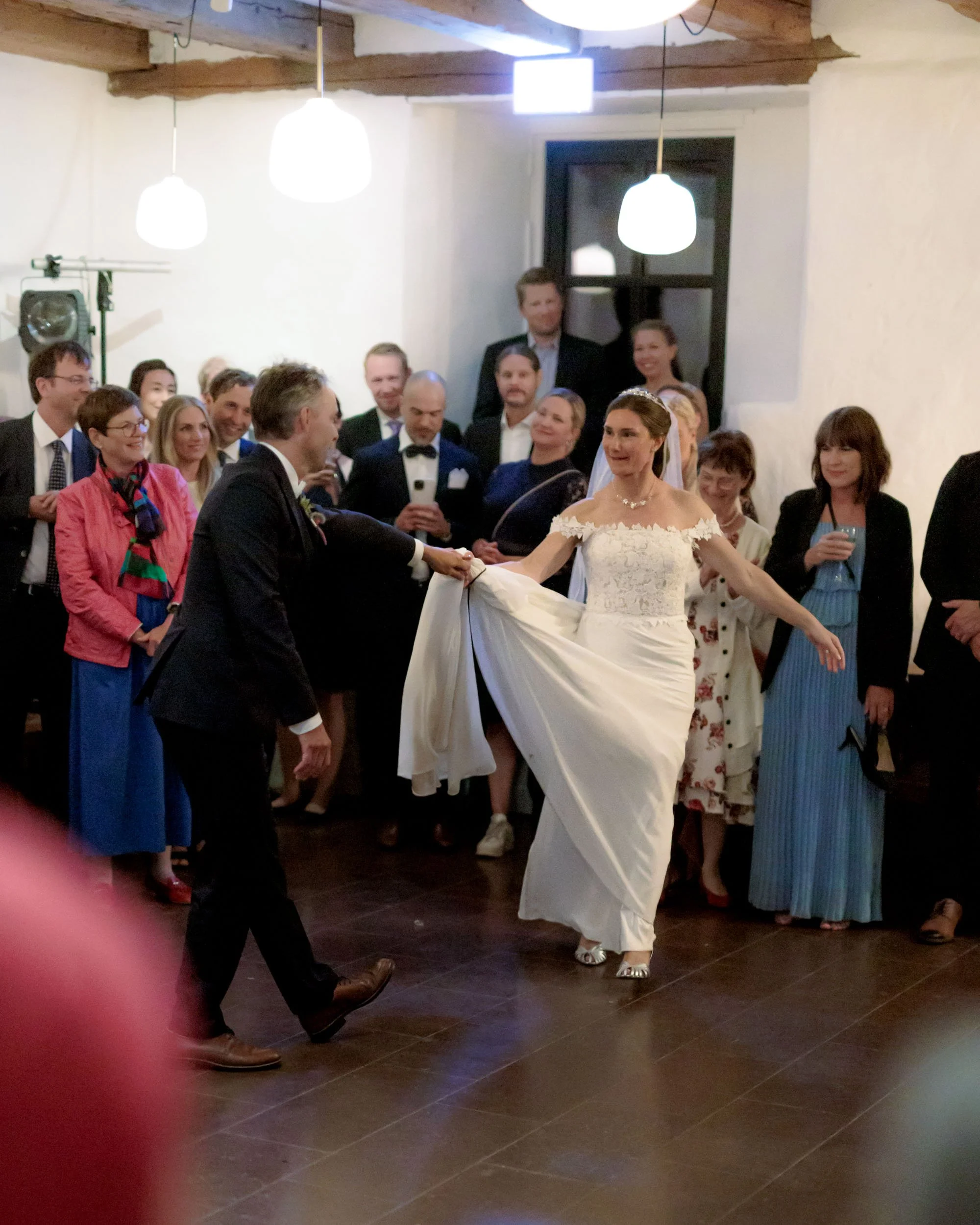 Brudparet dansar och håller varandra i händerna framför ett gäng gäster på en fest i ett rum med takbjälkar och vita väggar.