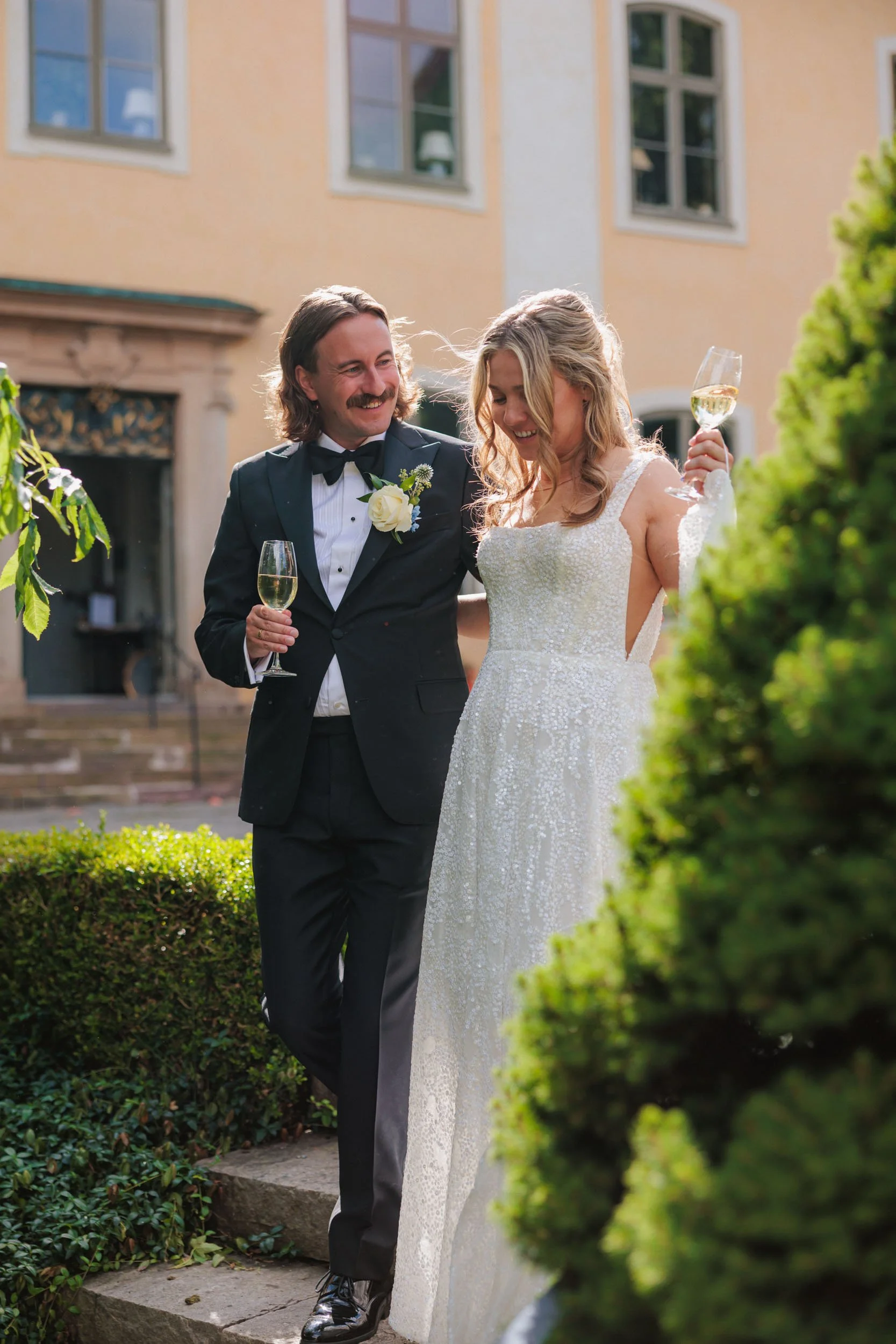En man och en kvinna i formella kläder som firar ett bröllop utomhus, båda håller glas med vitt vin och ler.