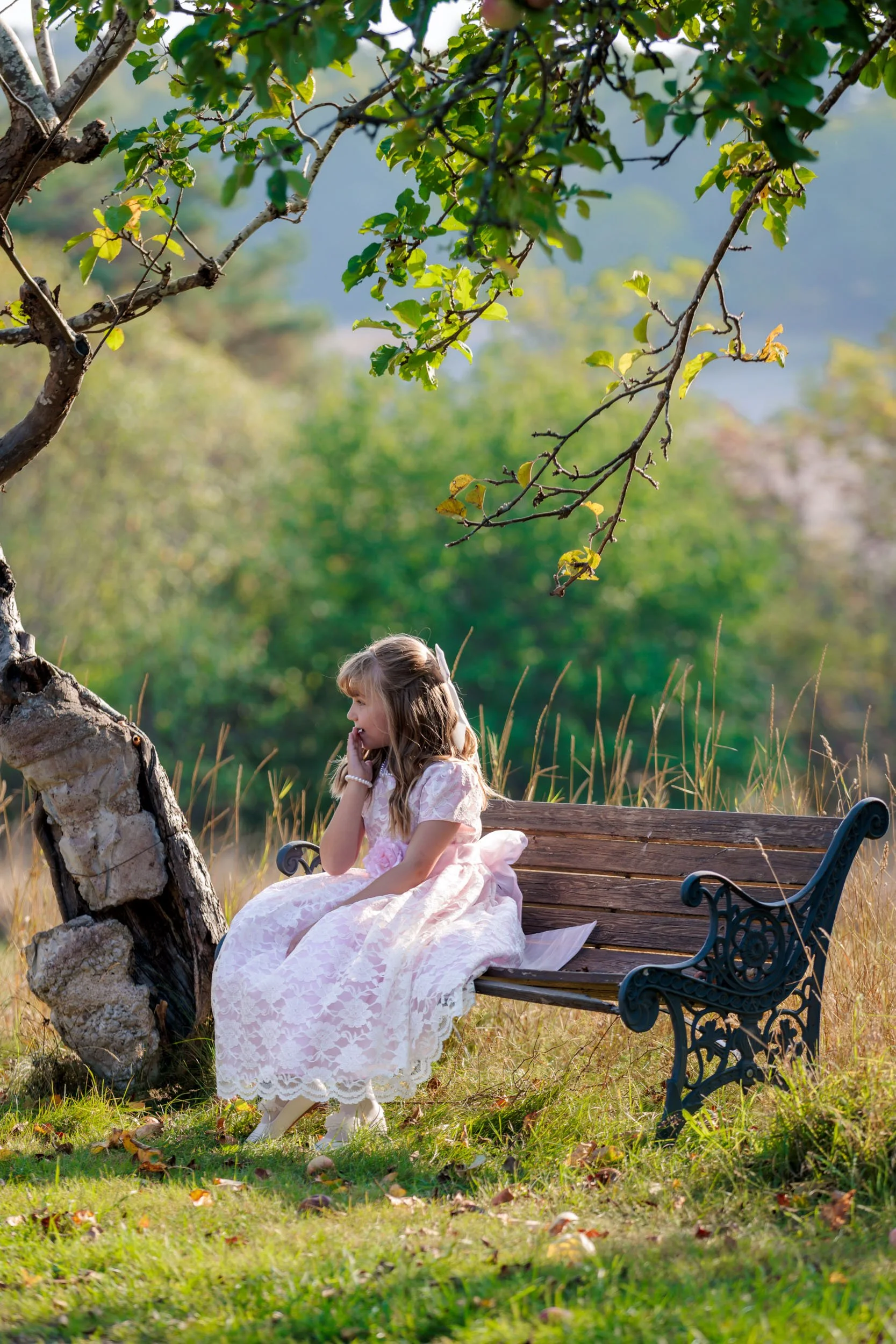En flicka i en vit spetsklänning sitter på en träbänk under ett träd i en grön park.