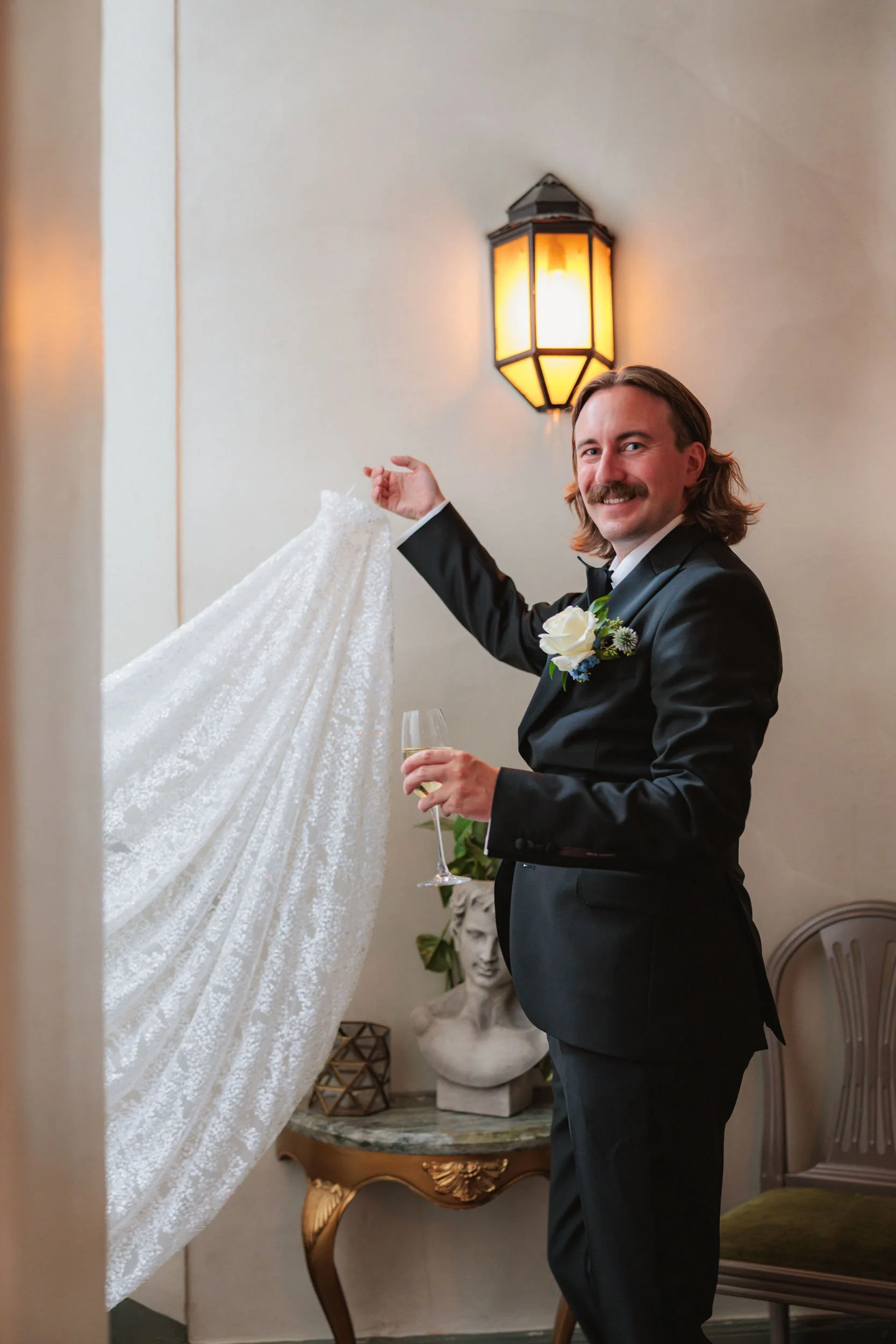 Man i smoking och med blommor i bröstet, som håller i ett glas champagne och en lakan i handen, står i ett rum med en vägglampa och konstverk.