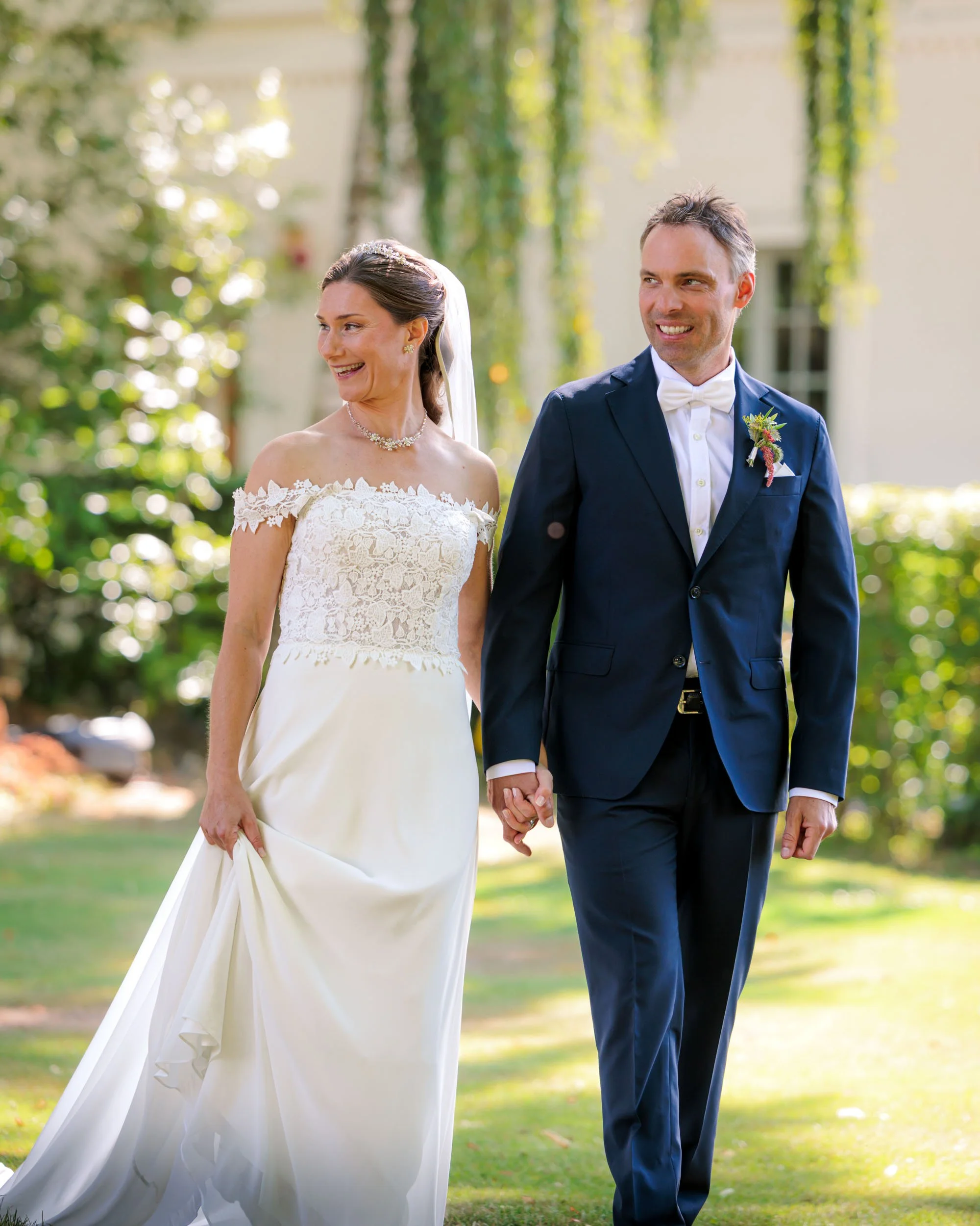 En brud och en brudgum håller handen och går utomhus på sin bröllopsdag, omgiven av grönt och träd, solen skiner.