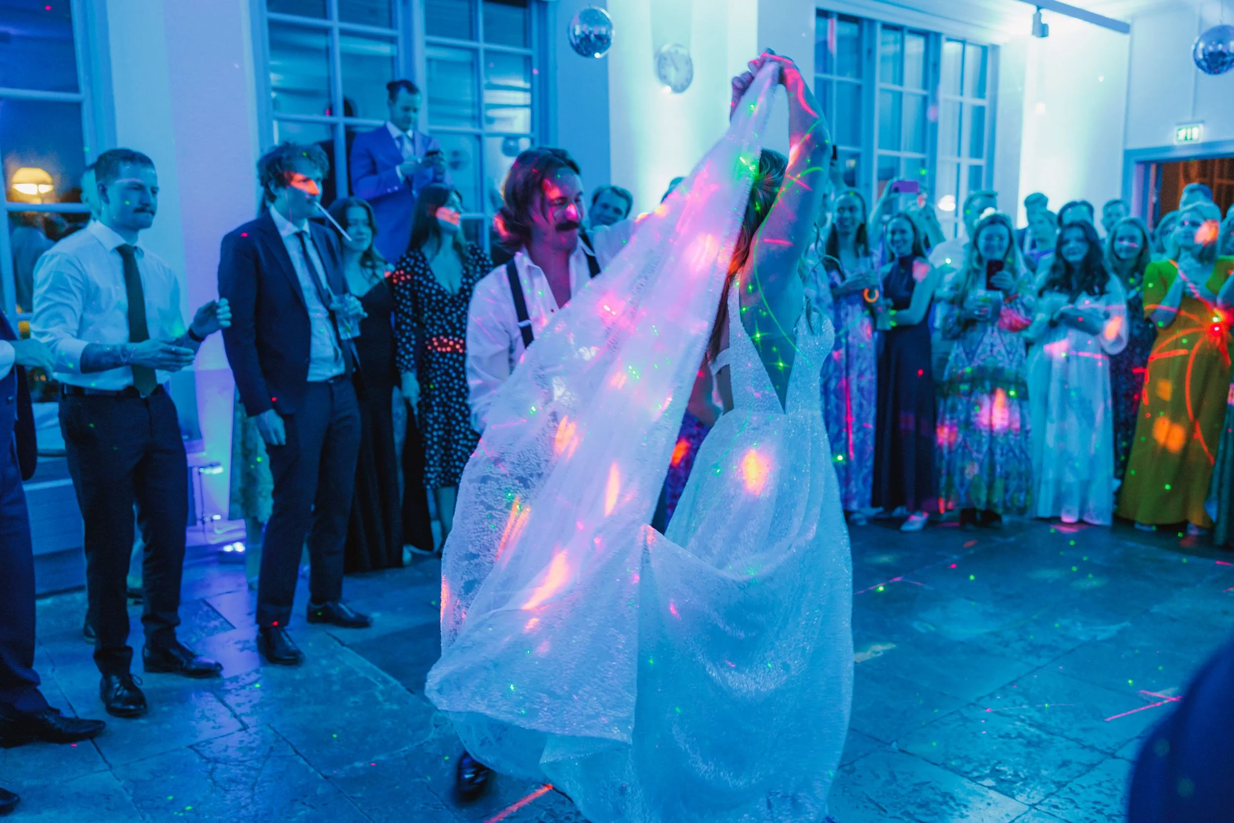 En person dansar med en tygduk i händerna under ett festligt evenemang, omgivet av gäster i formella kläder och färgglada klänningar.