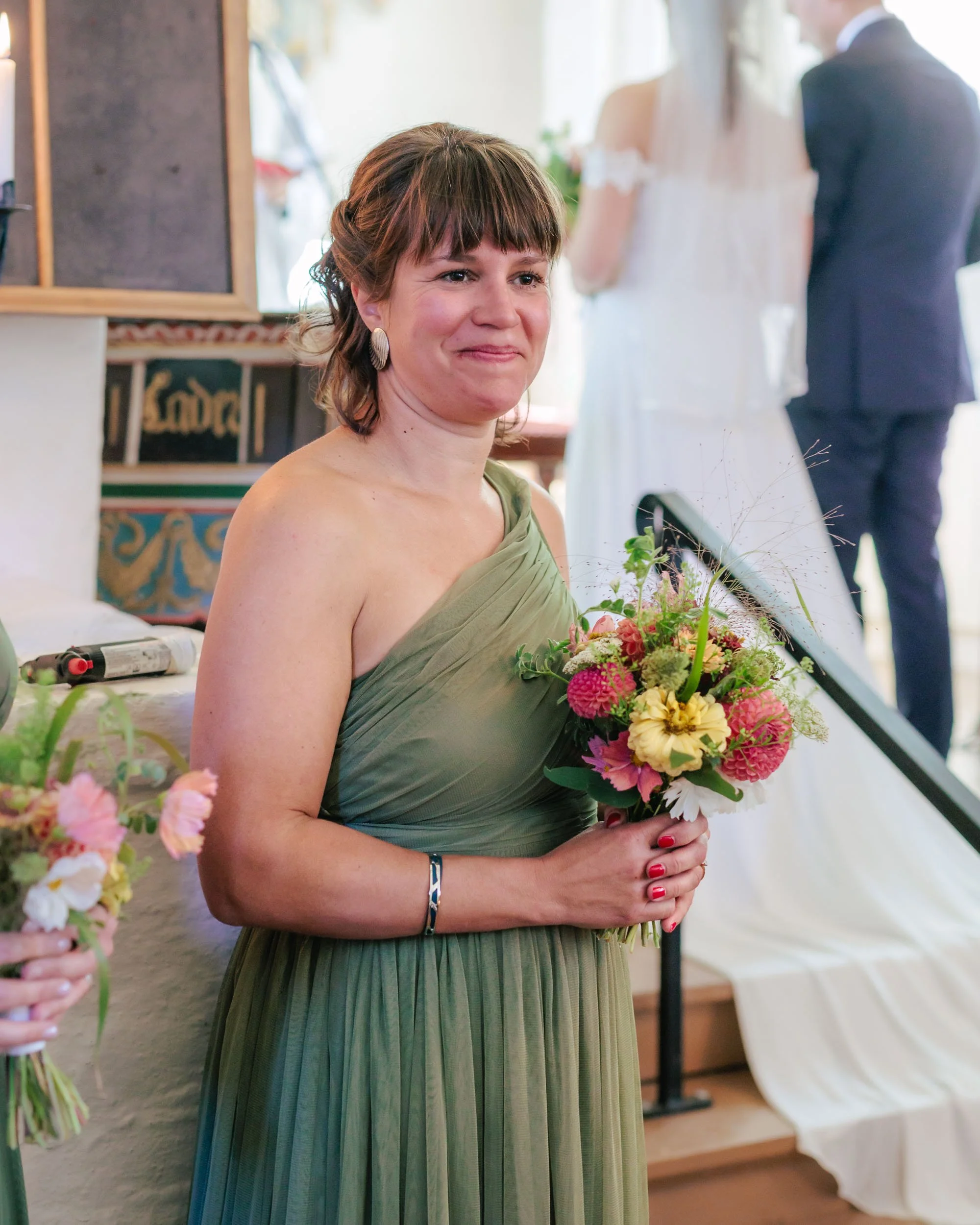 En kvinna i en grön klänning håller en bukett blommor och ler under ett bröllop.