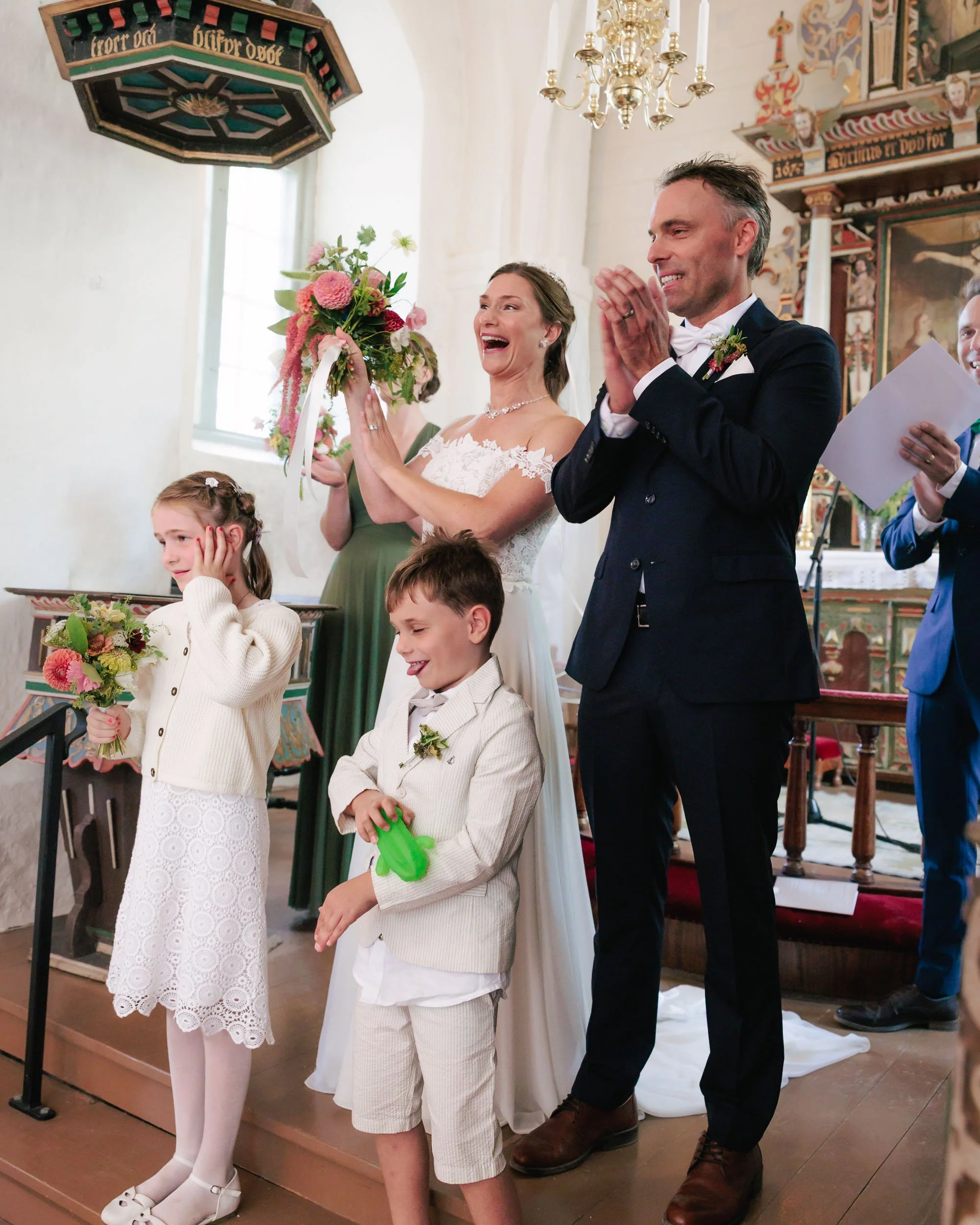 Brudparet och barn på en bröllopsceremoni i en kyrka, med leende föräldrar och barn, där barnen håller blommor.