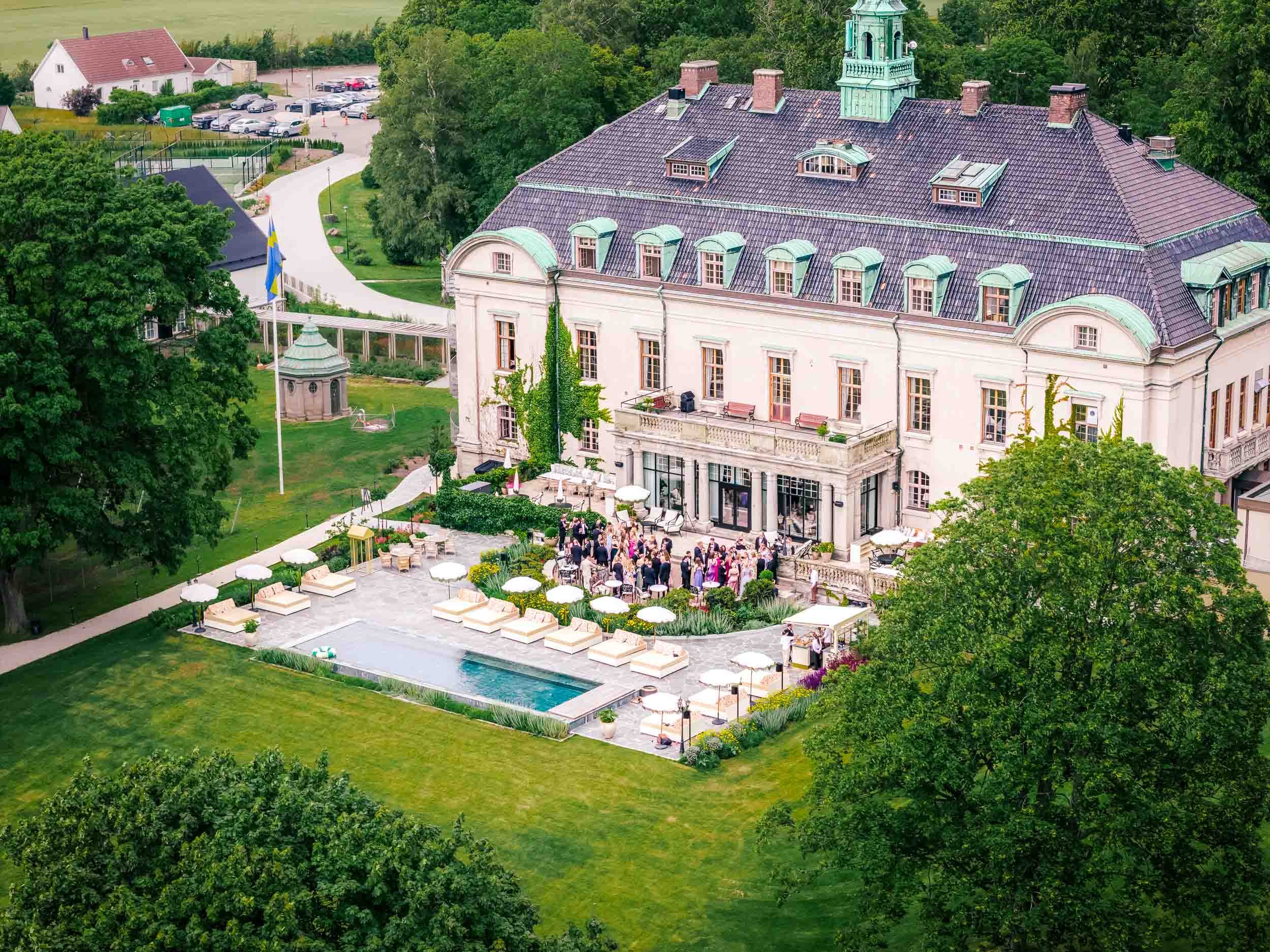 Festligheter utanför en stor vit herrgård med många gäster samlade på terrassen, omgivande grön trädgård och pool.