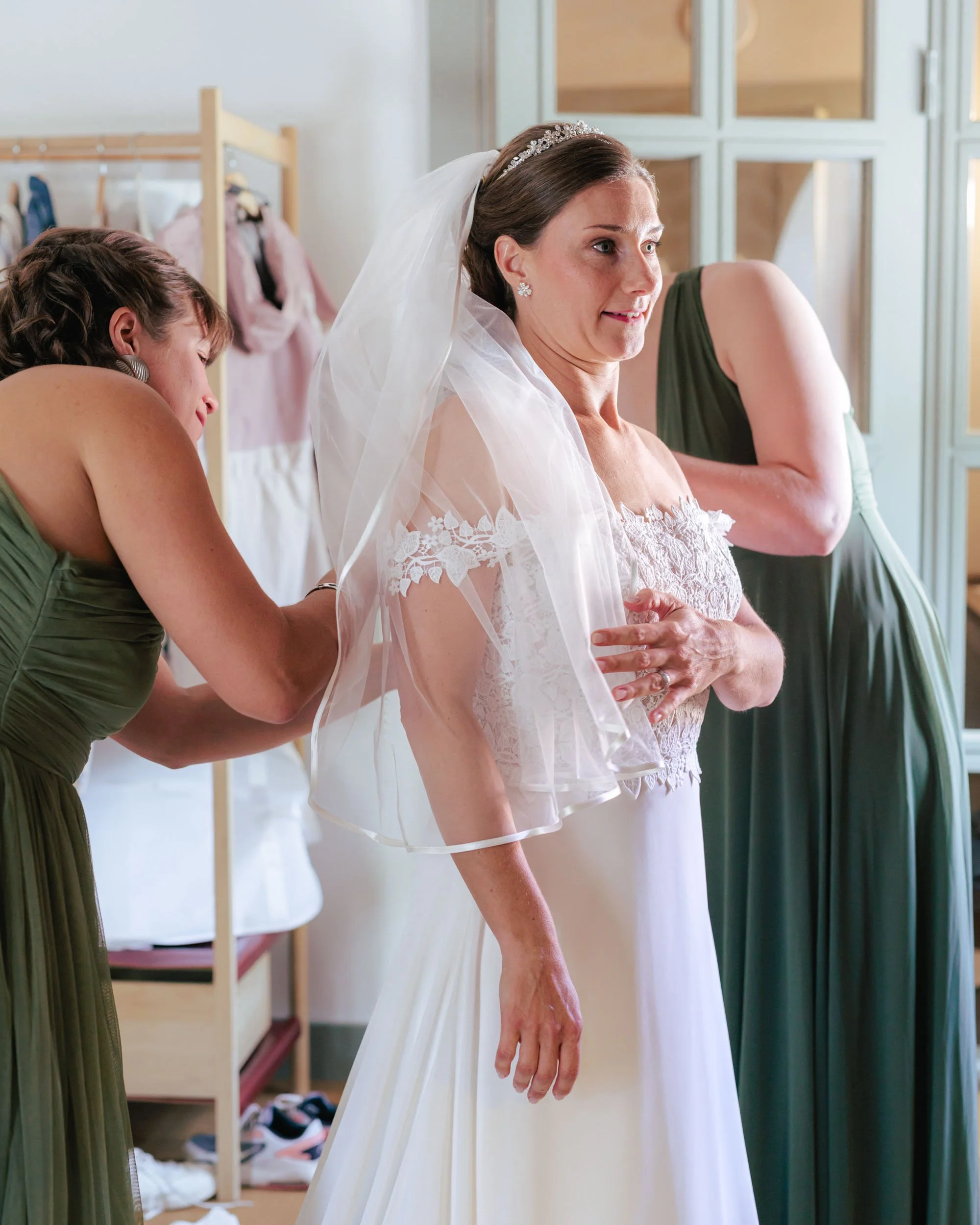 En brud klär på sig sin brudklänning medan två kvinnor hjälper henne, i ett rum med garderober och kläder i bakgrunden.