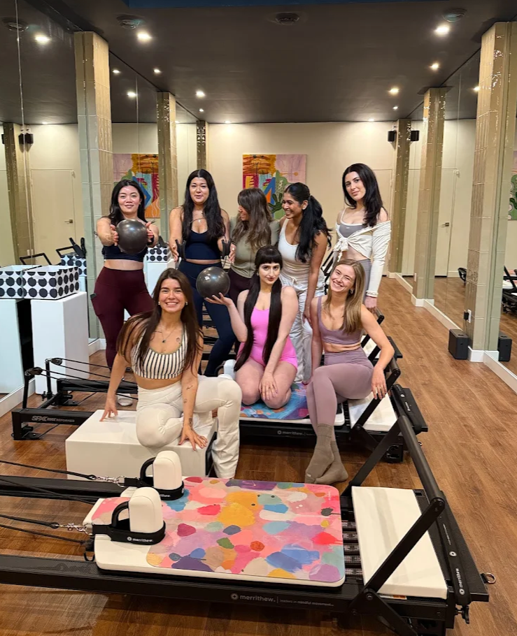 A group of 8 young woman, all weating workout sets, posing with Pilates balls and sitting on a reformer.