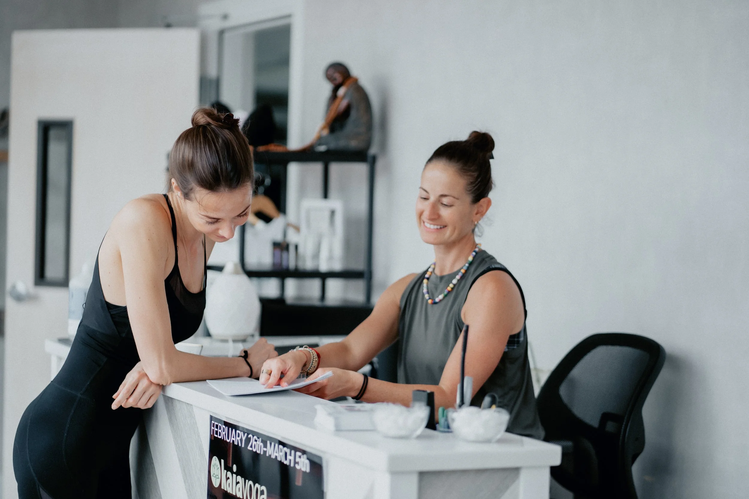A woman that's part of Kaia Yoga's team is at the front desk of the yoga studio explaining a list of written instructions to another woman, who's a new client at the boutique fitness studio.