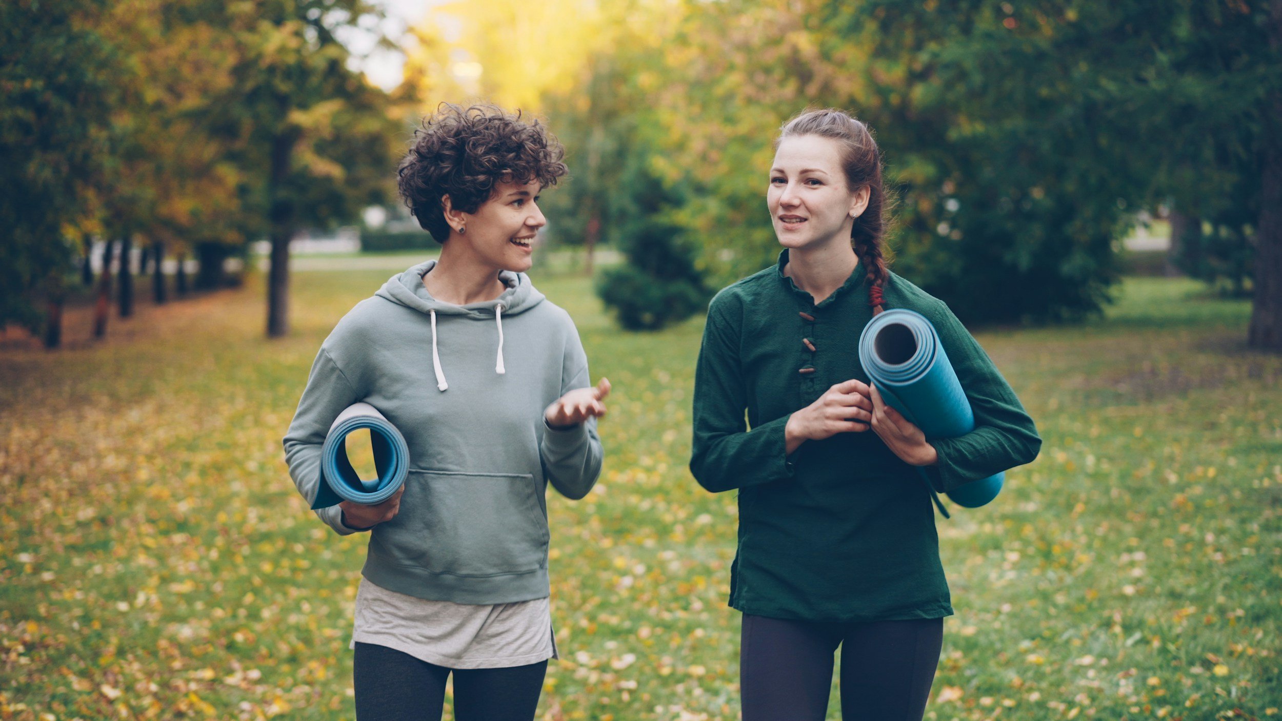 Two women with yoga mats talking in the park surrounded by trees.
