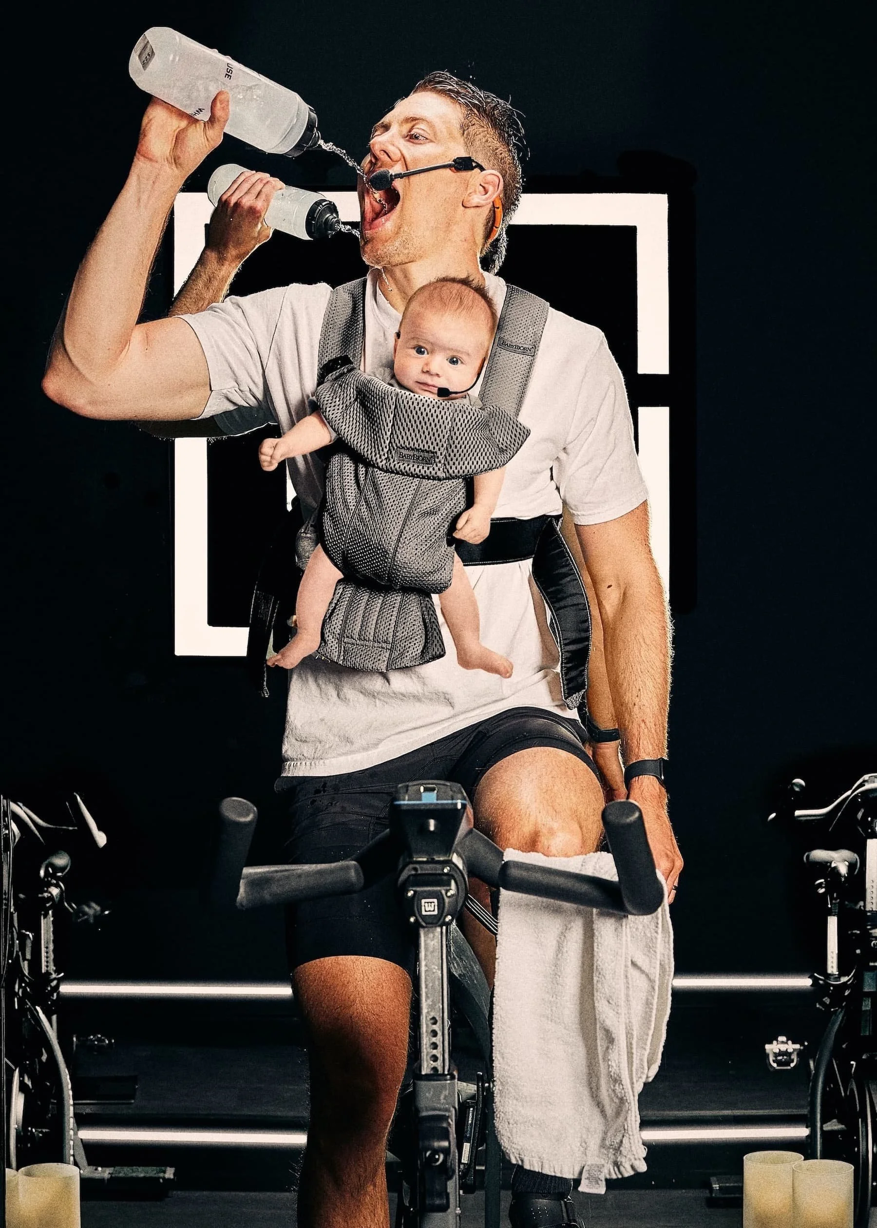 A man on an indoor bicycle drinks water with a baby strapped to his chest.