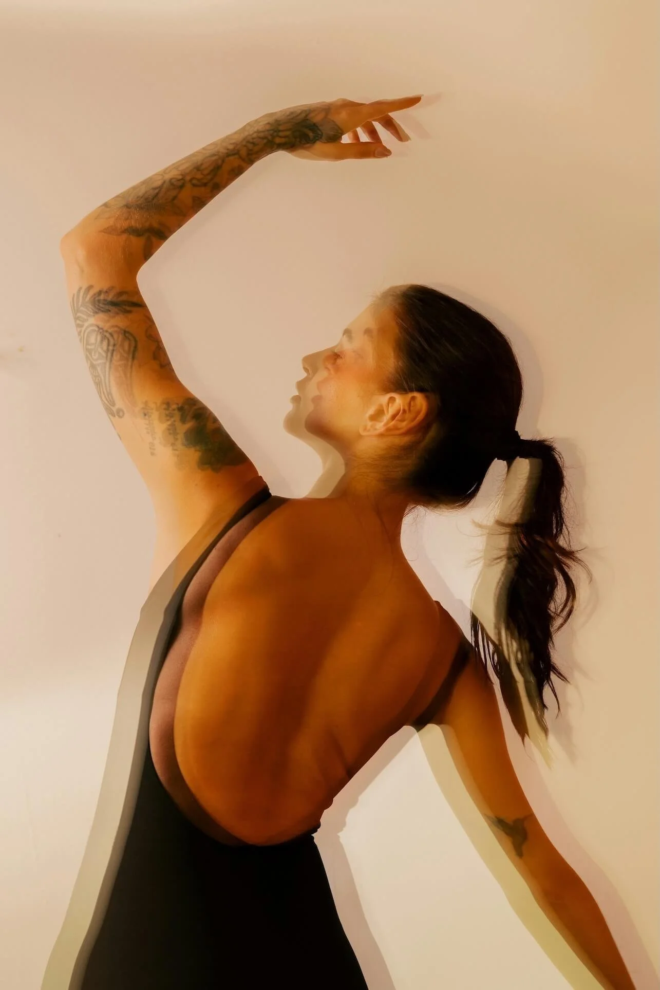 A woman with tattoos on her arm and side, wearing a black top, is stretching her arm upward and to the side, with her head turned upward and her long hair in a ponytail, against a plain white background.