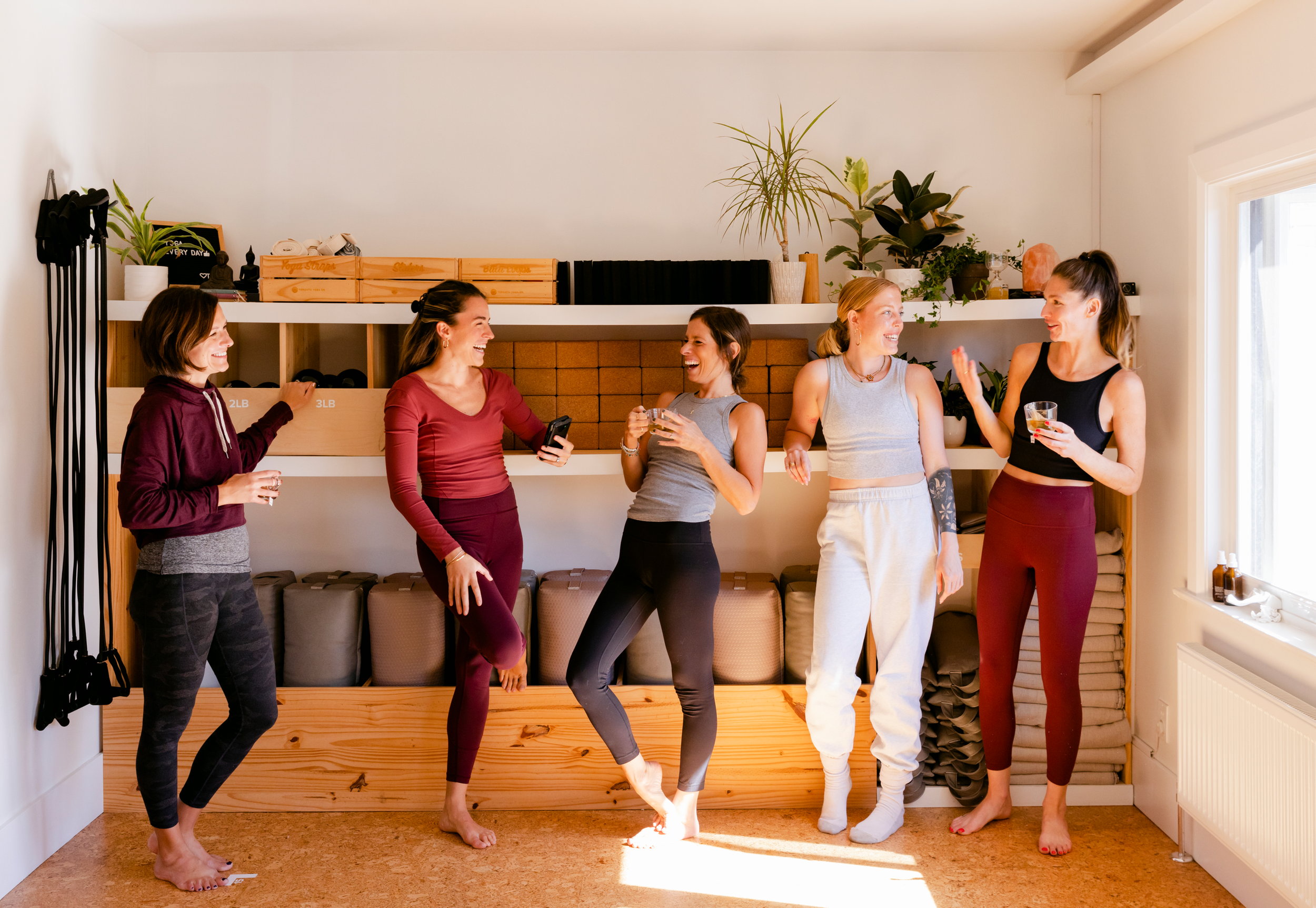 Five women standing barefoot in a line, engaging in conversation and laughter in a bright room with potted plants on a shelf and wooden storage boxes behind them.