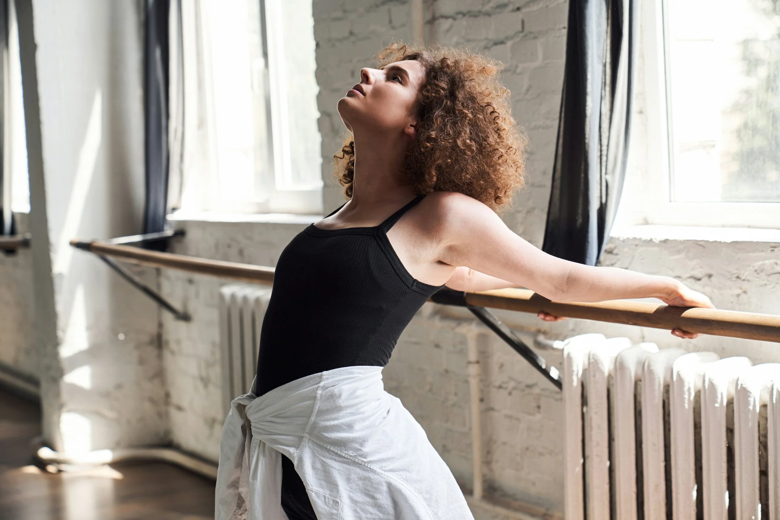 A woman with curly hair in a black sleeveless top and white pants stretching at a ballet barre in a dance studio with white brick walls and large windows.